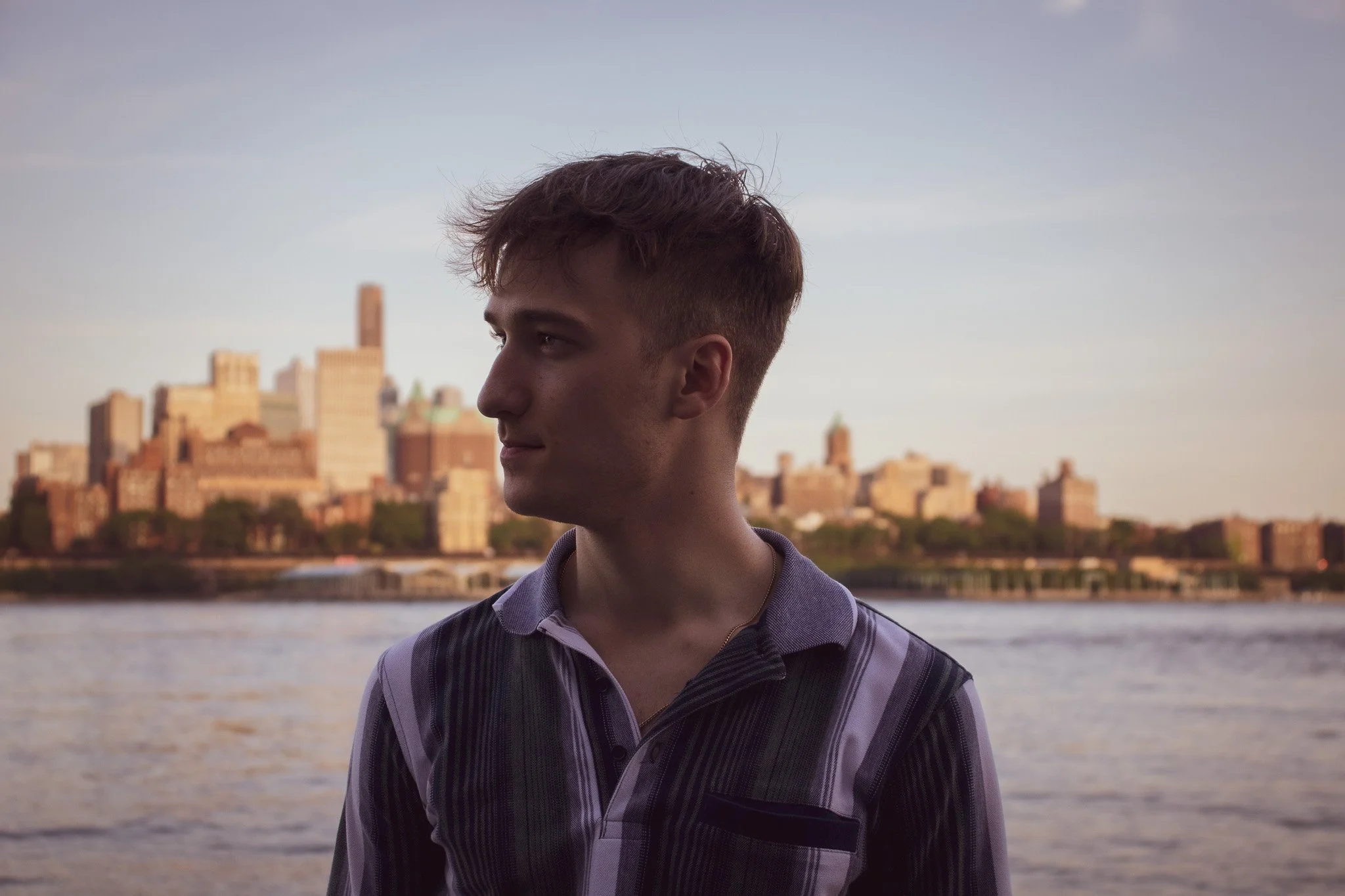 A young man with short, wavy hair, wearing a striped polo shirt, is standing outdoors by a river, with a city skyline in the background during sunset.