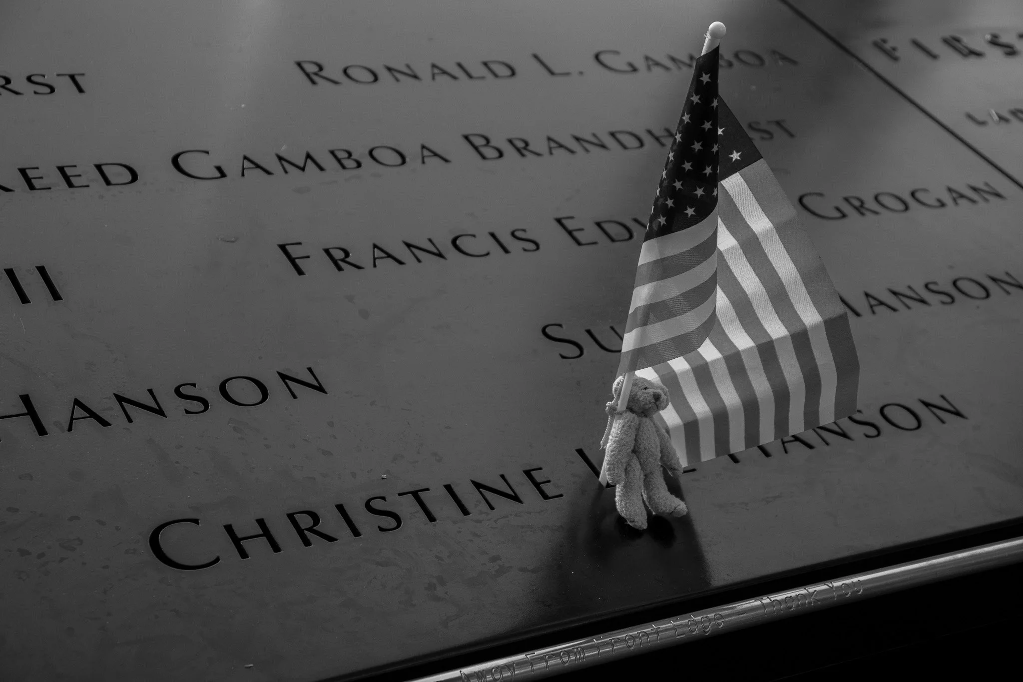 A small teddy bear holding an American flag is placed next to a memorial plaque on the ground. The plaque has engraved names of individuals, including Ronald L. Gamboa and Francis Edmison, and other names are partially visible.