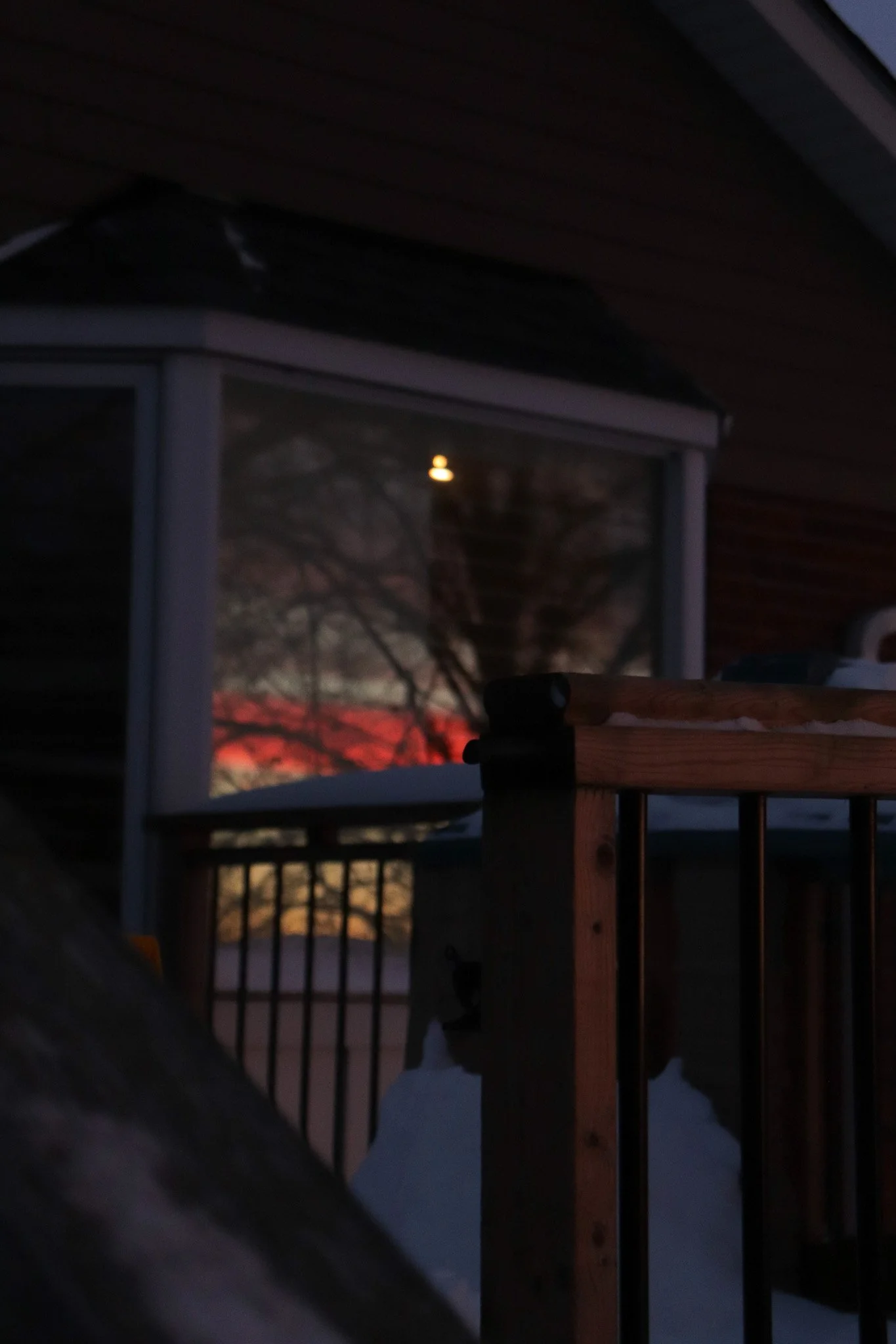 Nighttime outside view of a house porch with a window reflecting a tree and the moon, wooden railing, and ceiling.