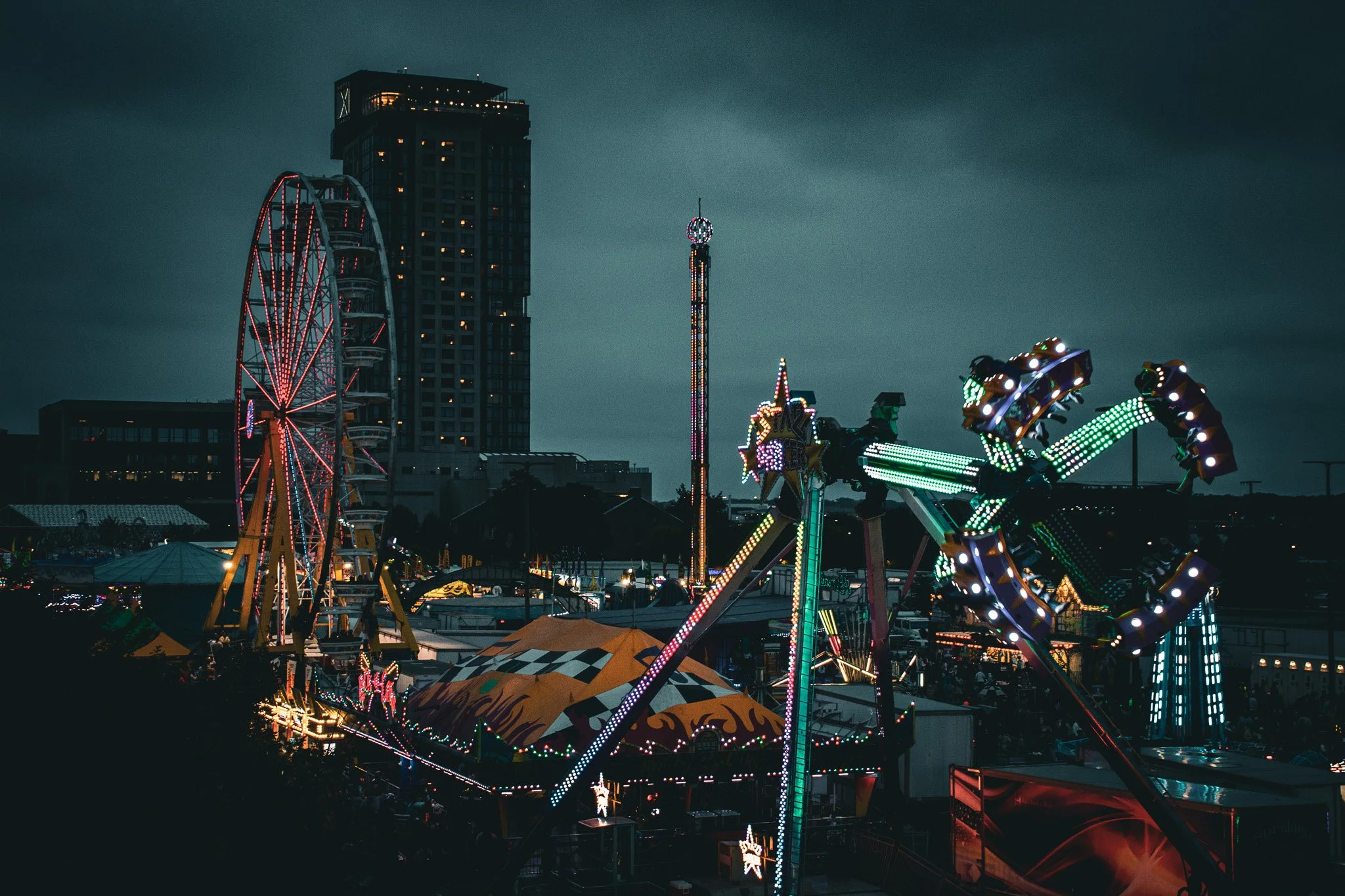 Nighttime view of a carnival with illuminated rides, including a Ferris wheel and spinning rides, against a dark cloudy sky with tall buildings in the background.
