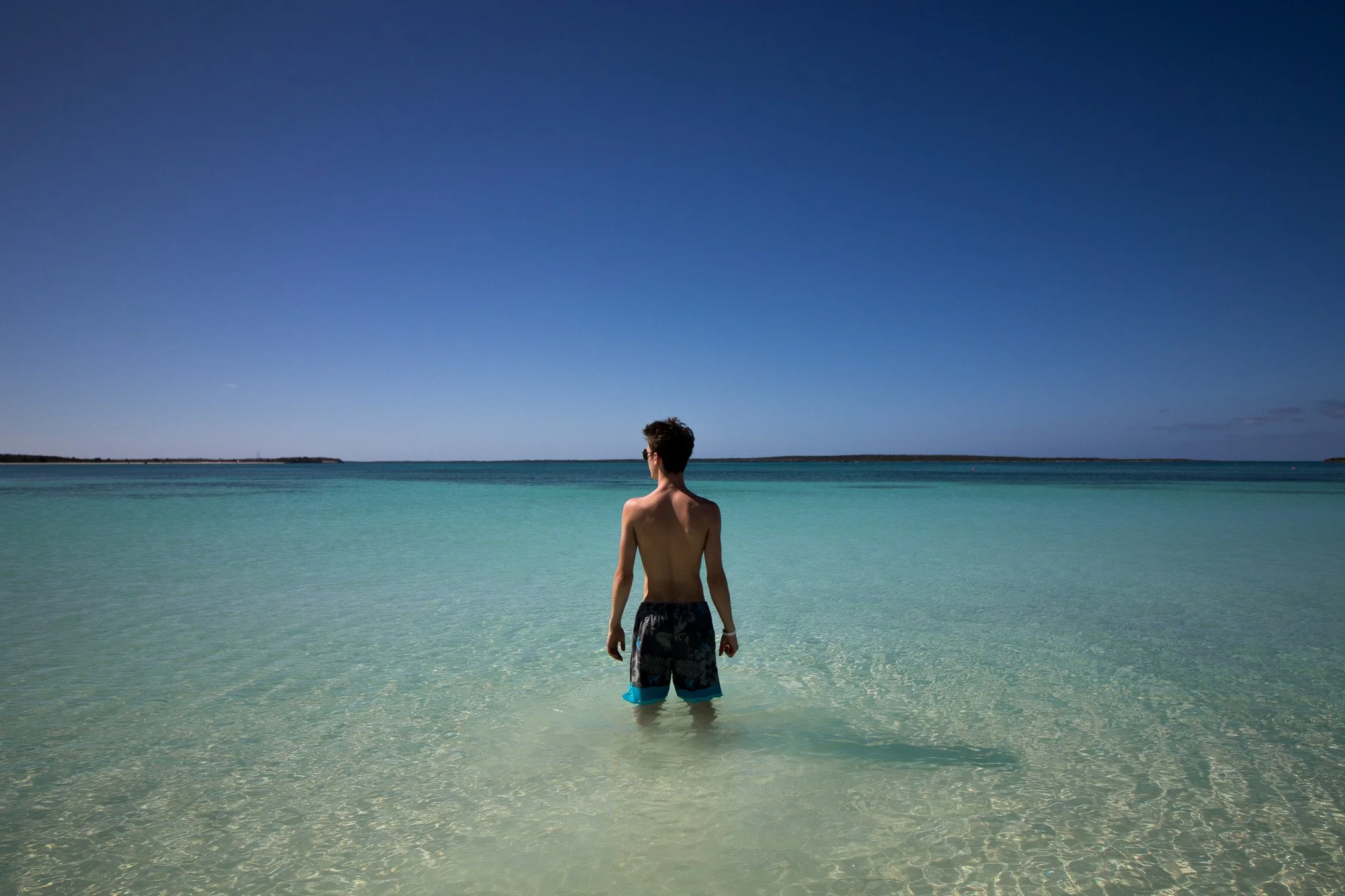 A person with short dark hair wearing swim trunks stands in shallow clear blue-green water at a beach, looking towards the horizon under a bright blue sky with a few clouds.