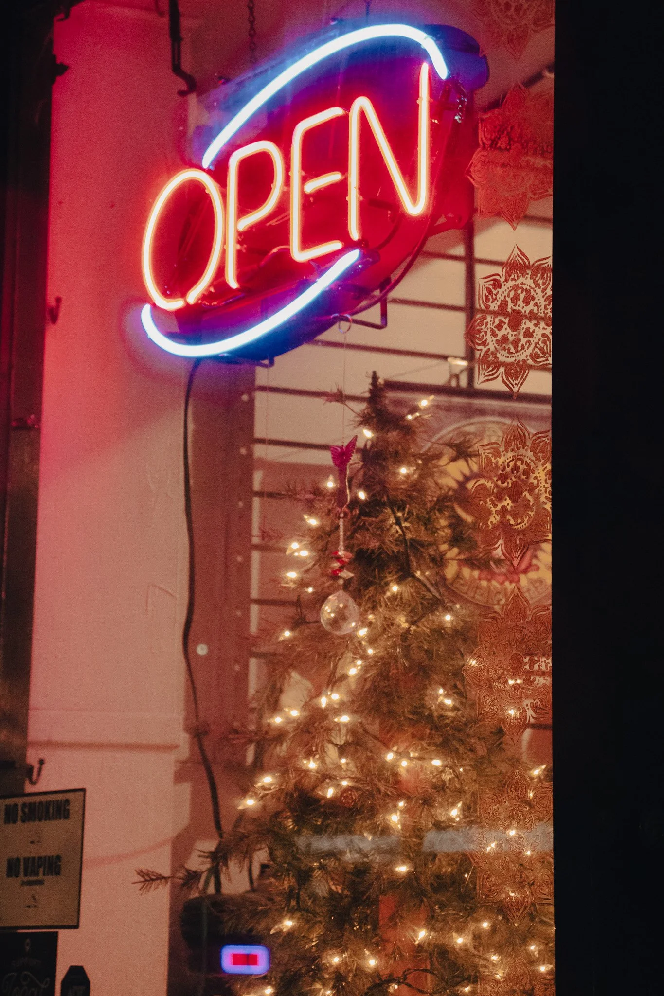 Neon sign that says 'OPEN' in red and blue colors hanging above a decorated Christmas tree with lights and ornaments, inside a room with patterned wallpaper and various signs on the wall.