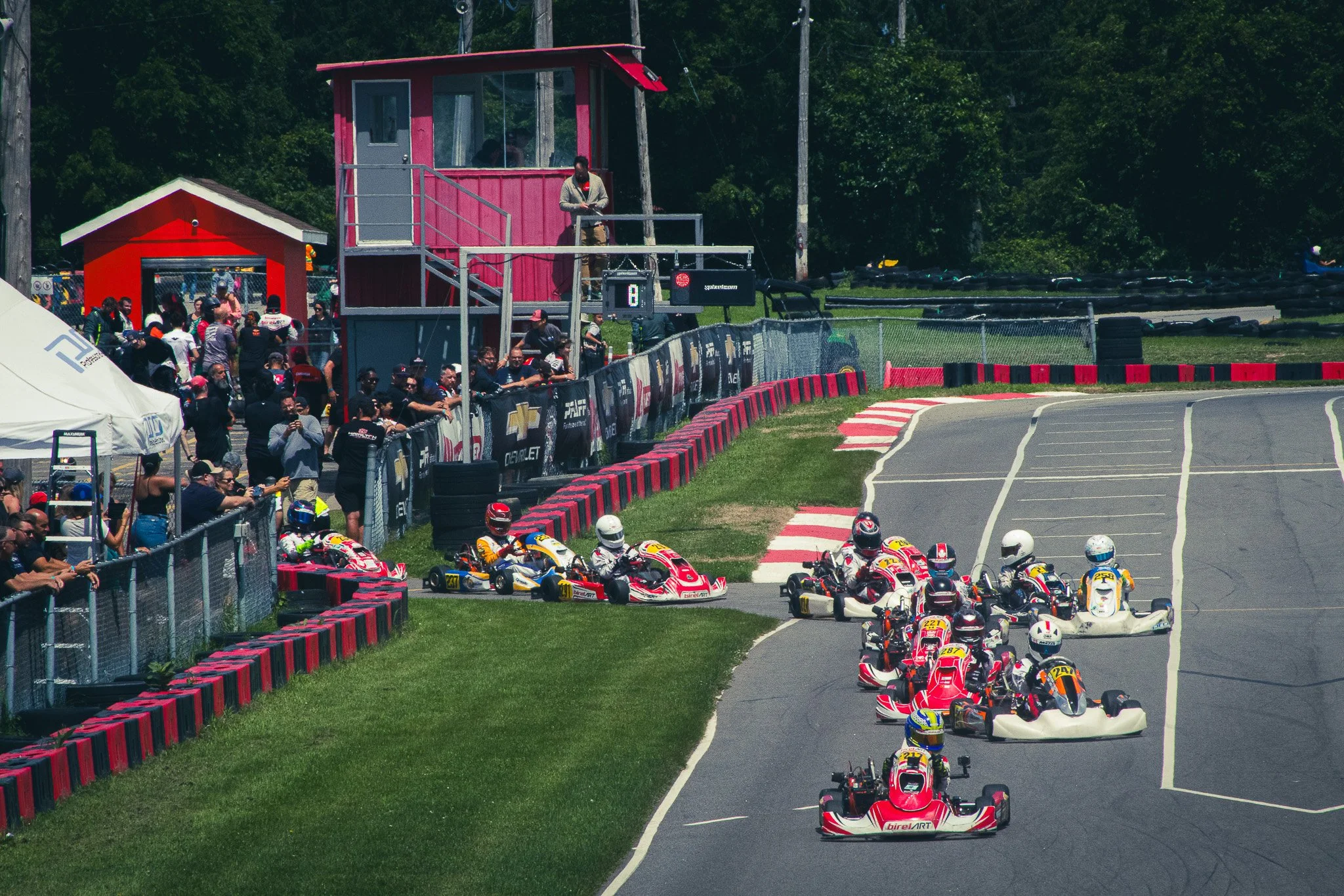 A go-kart race track with many go-karts lined up before a race, spectators watching from the side, and a pink control tower with an official standing on a platform. Trees are visible in the background.