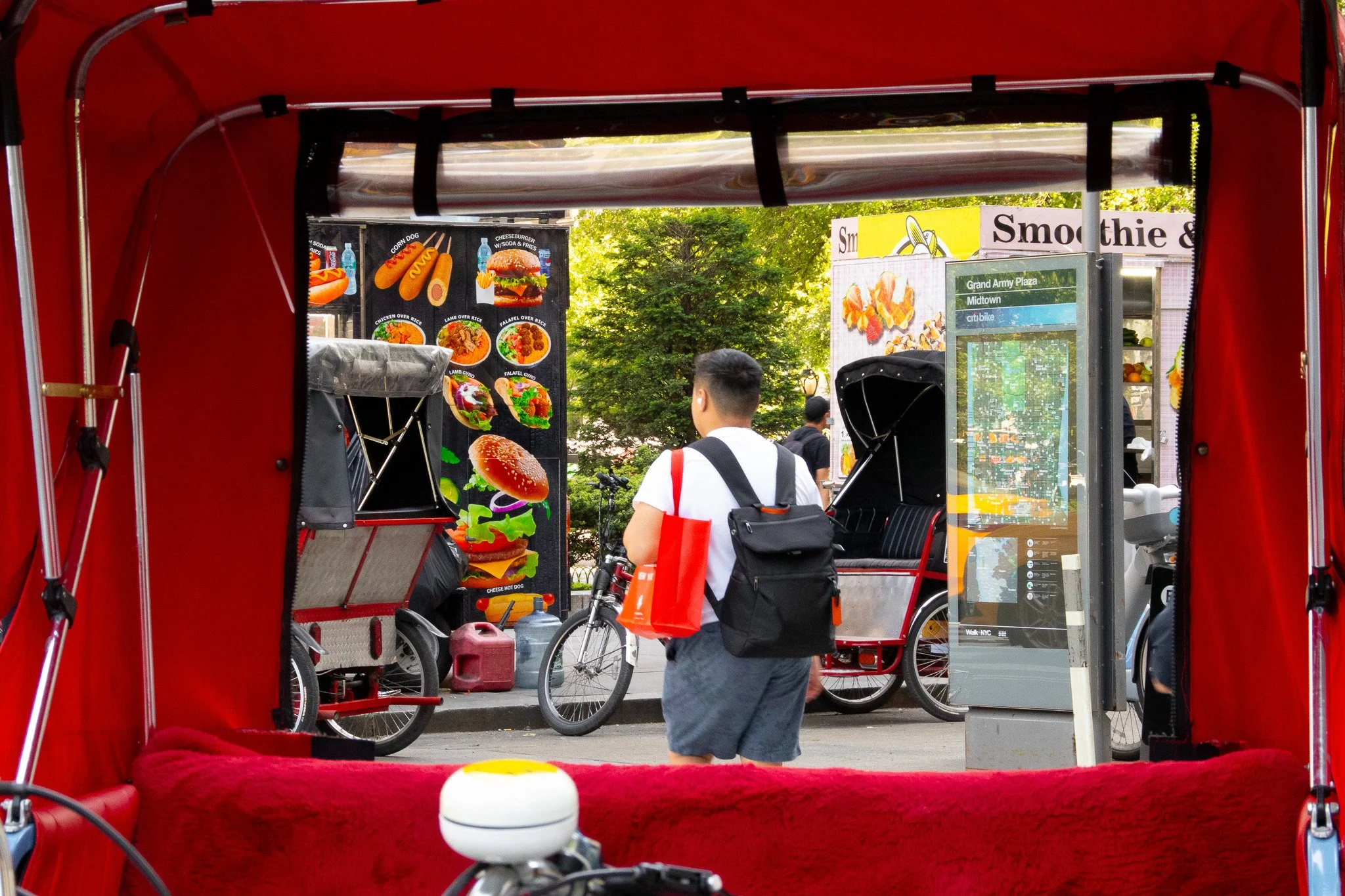 View from inside a red rickshaw looking out towards a street scene with people, bicycles, and signage for food and smoothies.