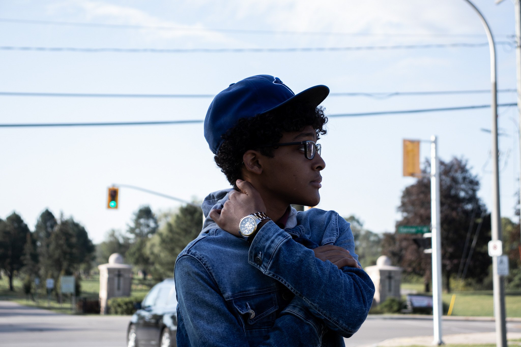 A young person with curly hair, glasses, and a denim jacket touching their shoulder, standing outdoors near a street with traffic lights and power lines, during daytime.
