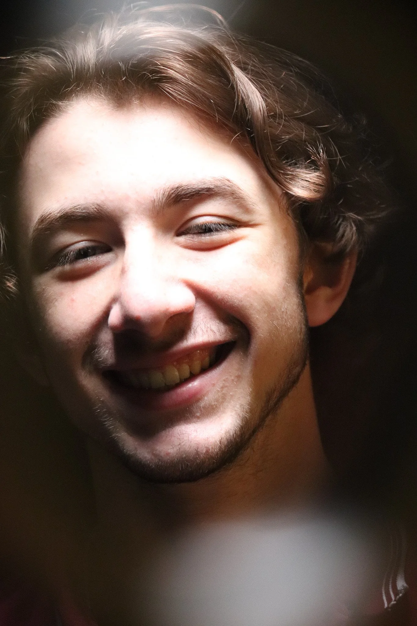 Close-up of a young man smiling with partially closed eyes and a dark background.