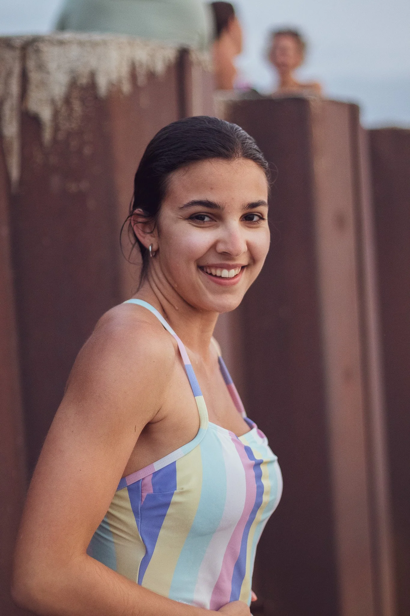 A woman with dark hair styled back wearing a colorful striped tank top, smiling at the camera with a brown wall in the background.
