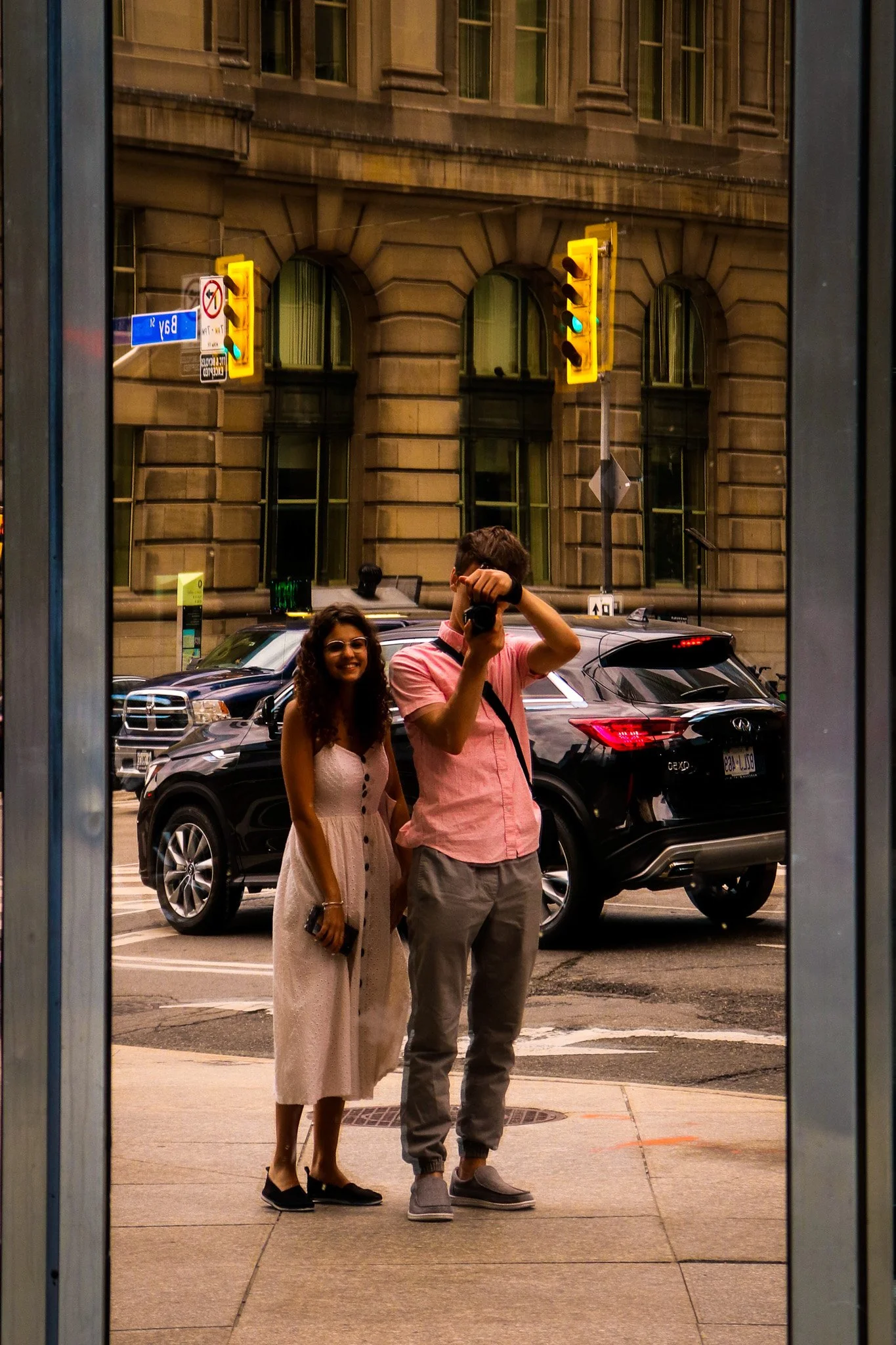 Reflection of a man and a woman taking a selfie on the sidewalk, with a city street scene and a building with arched windows in the background.