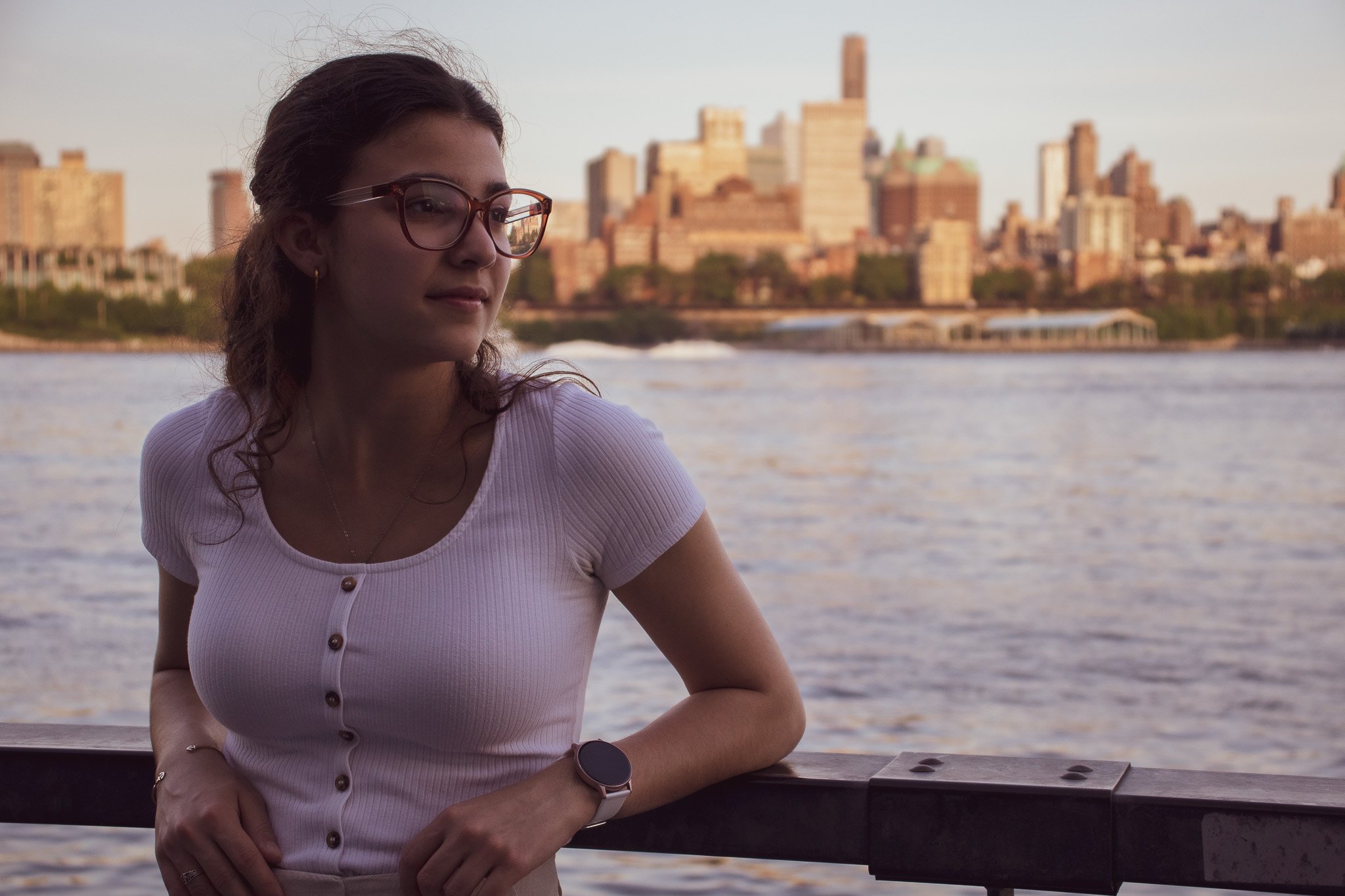 A young woman wearing glasses and a pink top with buttons, standing near water with a city skyline in the background.