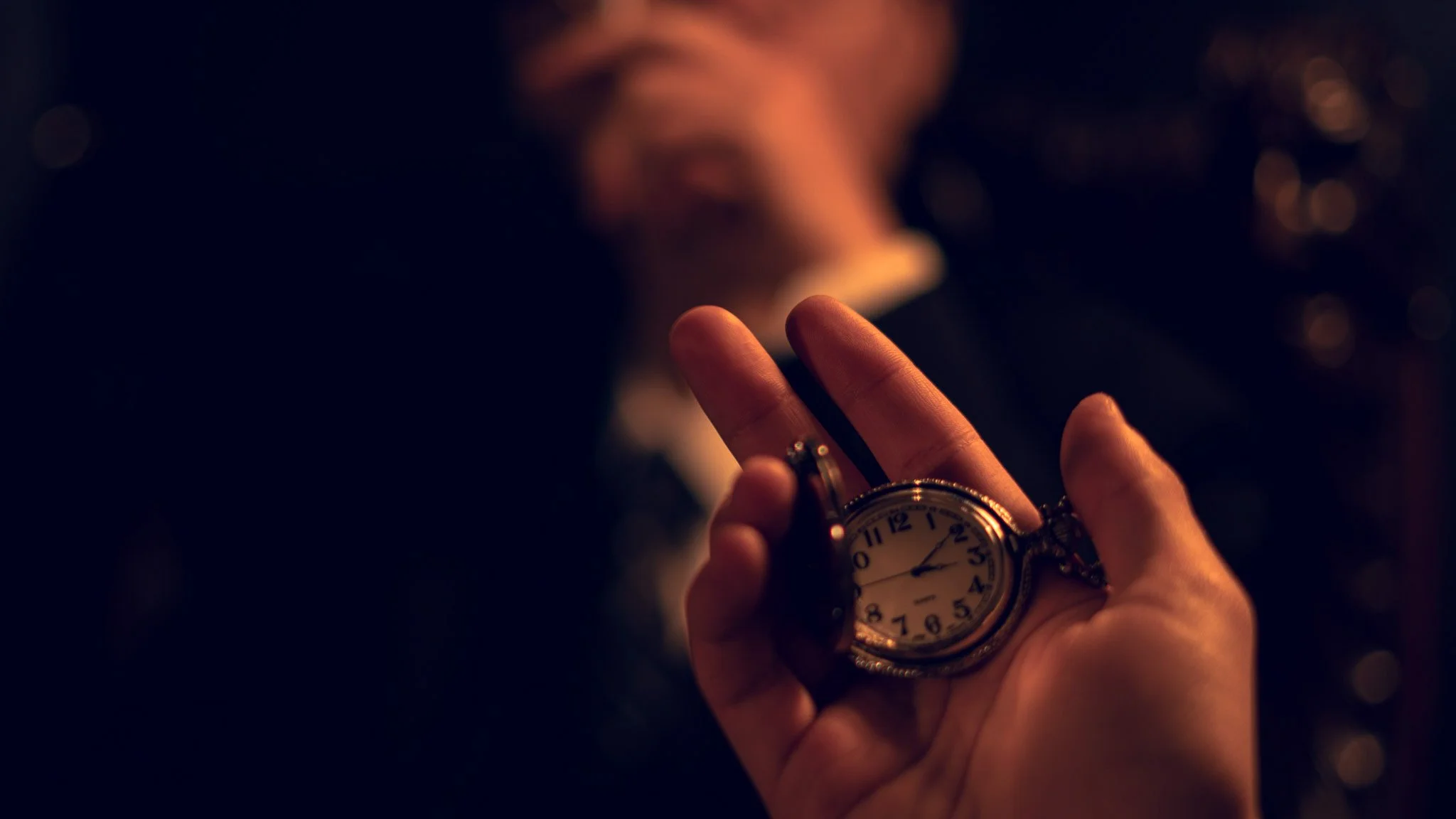 A person holding a pocket watch, with the focus on the watch and fingers, in a dimly lit setting.