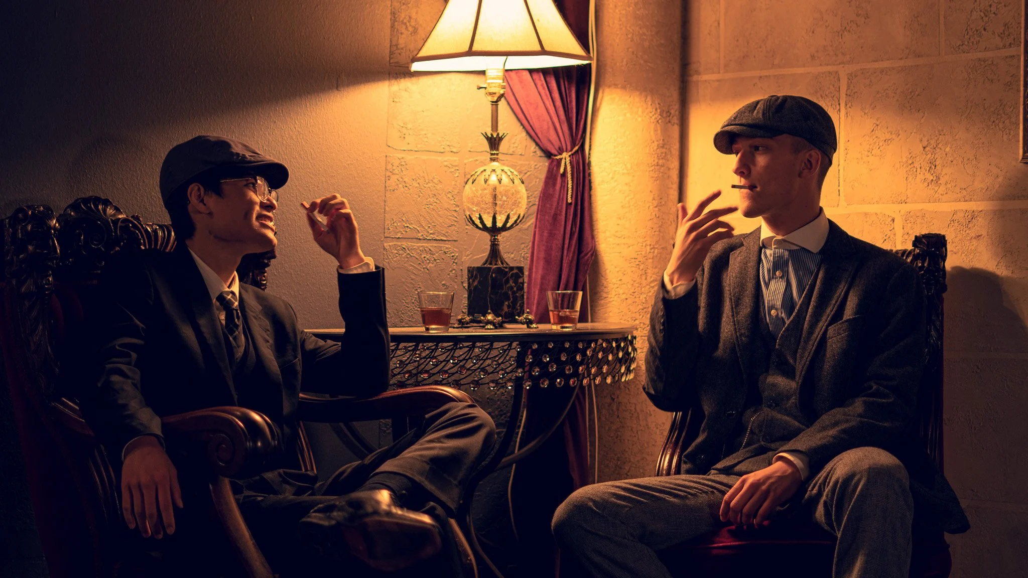 Two men in suits and flat caps sitting in a dimly lit room with vintage decor. One man is smoking a cigarette and the other is smiling, with a table and a decorative lamp between them.