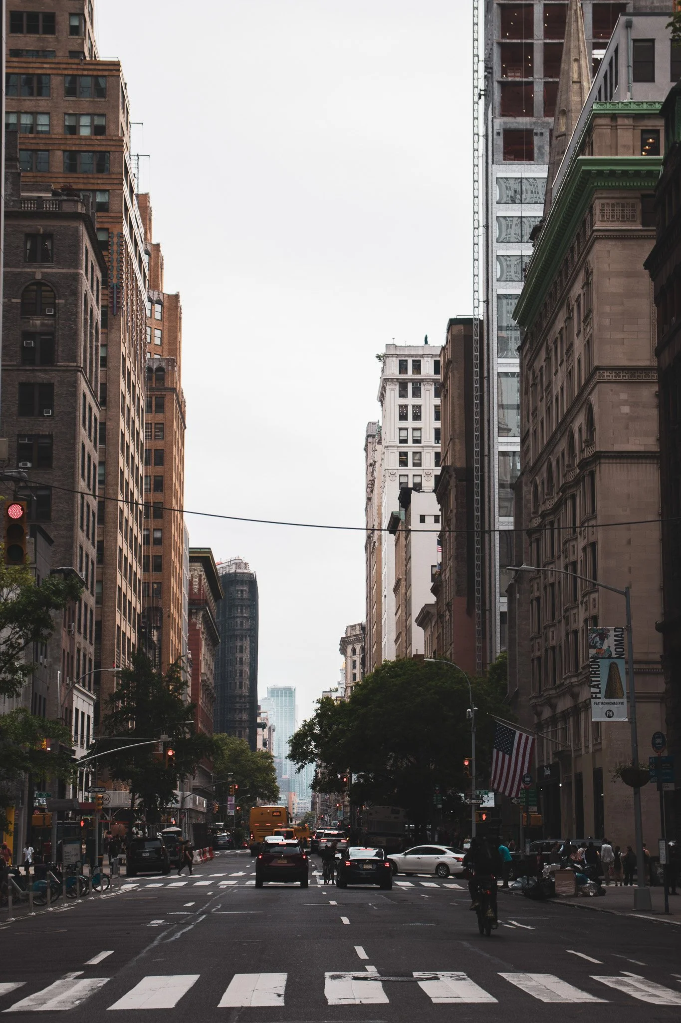 City street scene with tall buildings lining both sides, some construction, traffic, pedestrians, and trees visible, under an overcast sky.