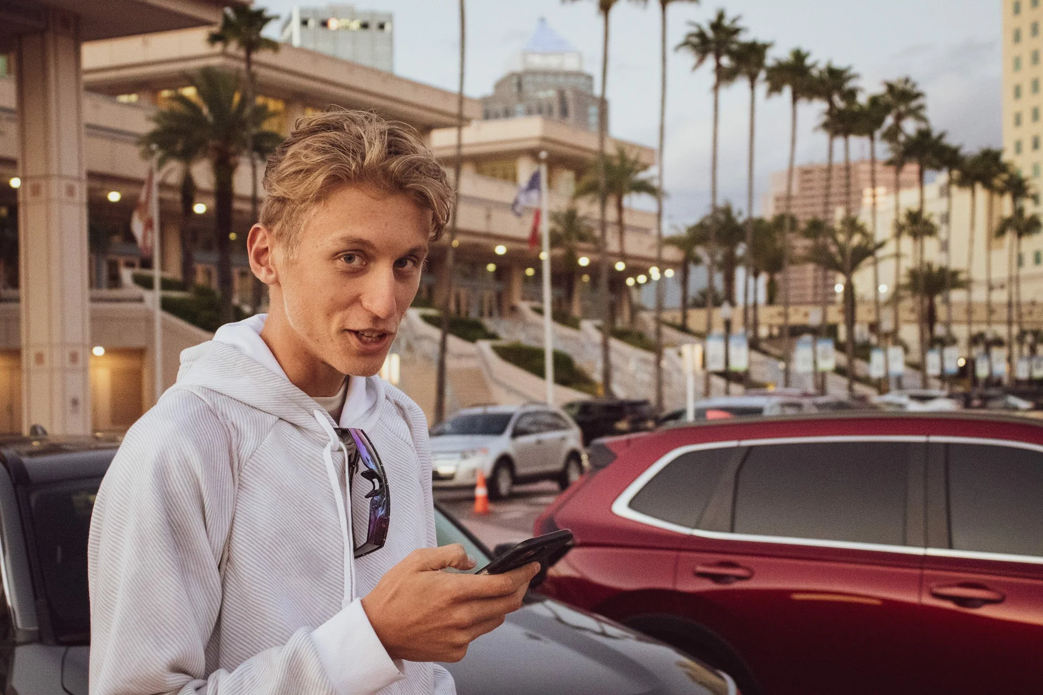 A young man with blond hair wearing a white hoodie and sunglasses hanging from his hoodie is standing outdoors, looking at a smartphone. Behind him are parked cars and a building with multiple floors, palm trees, and a city skyline in the background 