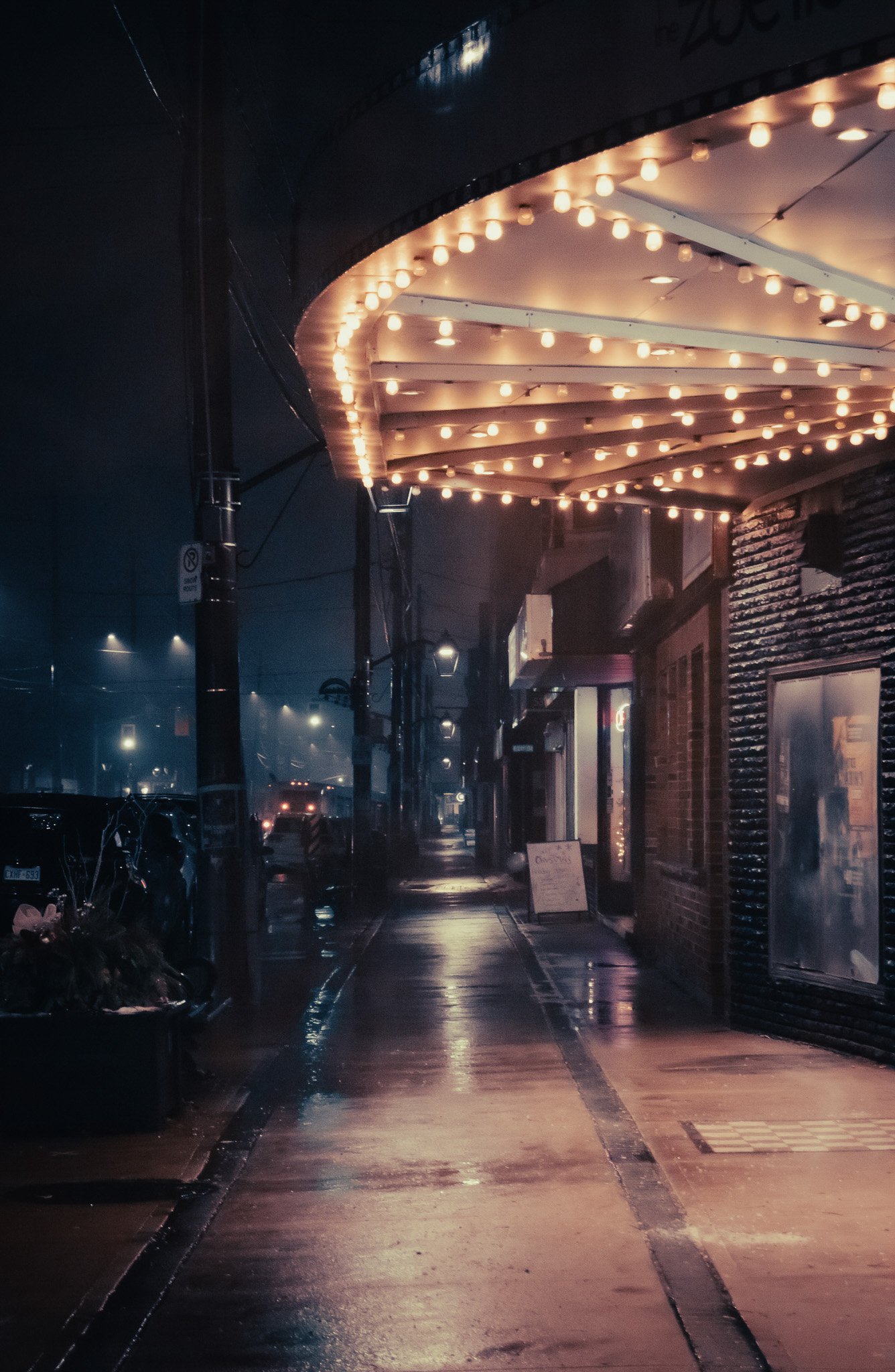 Nighttime street scene with wet pavement, illuminated by string lights under an awning, storefronts, parked cars, and a bike, all reflecting the glow of the lights