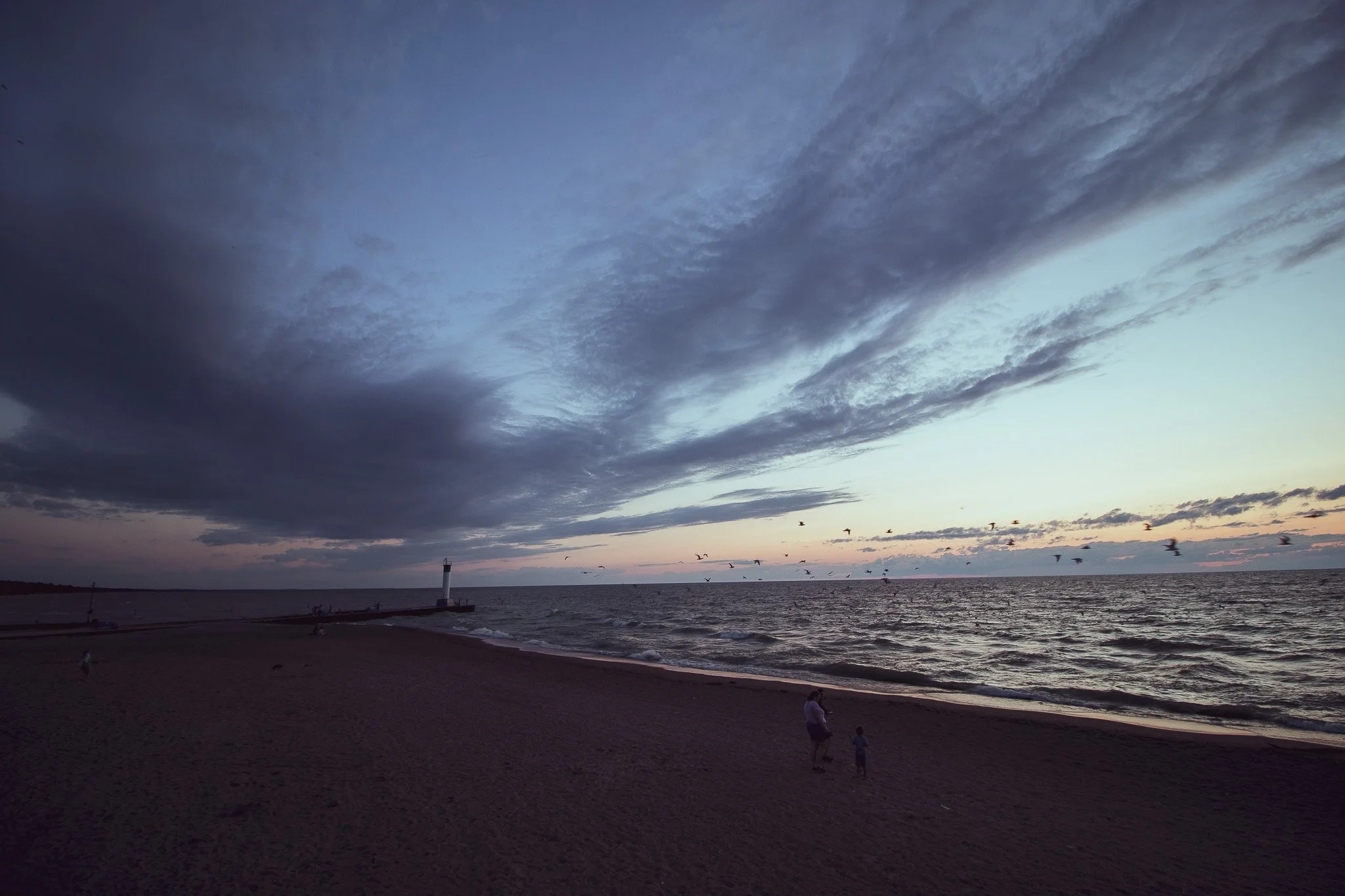 A beach at sunset with a cloudy sky, a lighthouse near the shoreline, and seagulls flying over the water, with people walking on the sand.