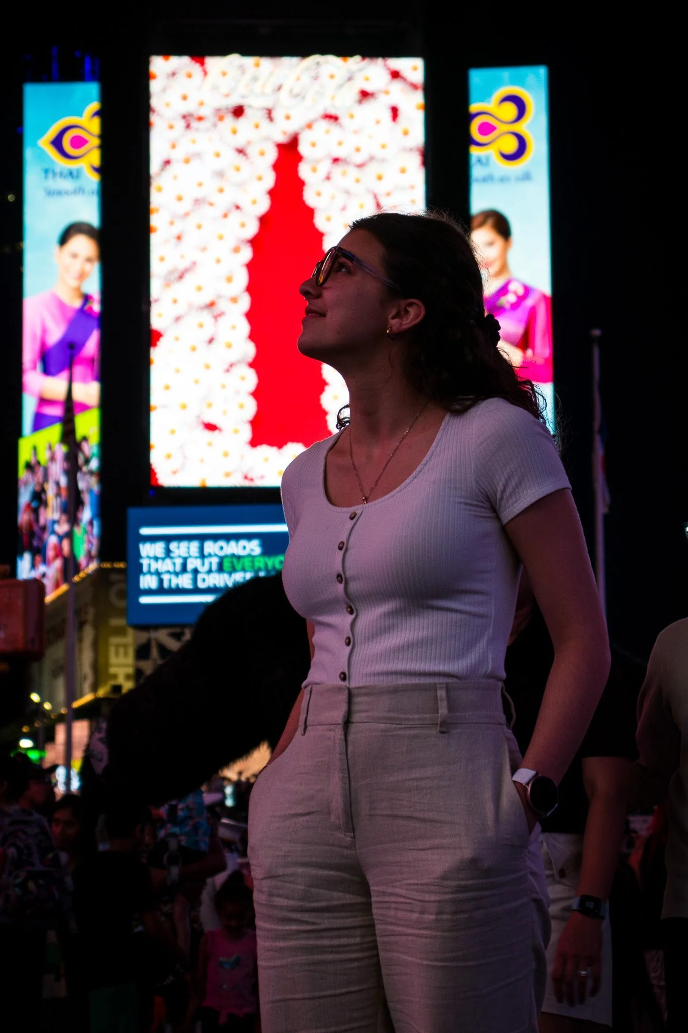 A woman with glasses and a white button-up shirt stands in a crowded outdoor area at night, illuminated by bright digital billboards and advertisements in the background.