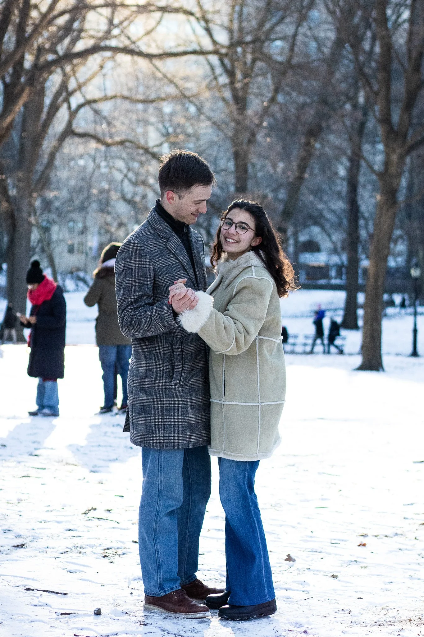 A young couple, a man and a woman, are holding hands and dancing outdoors on a snowy day in a park with leafless trees in the background. Other people are visible in the background enjoying the winter day.