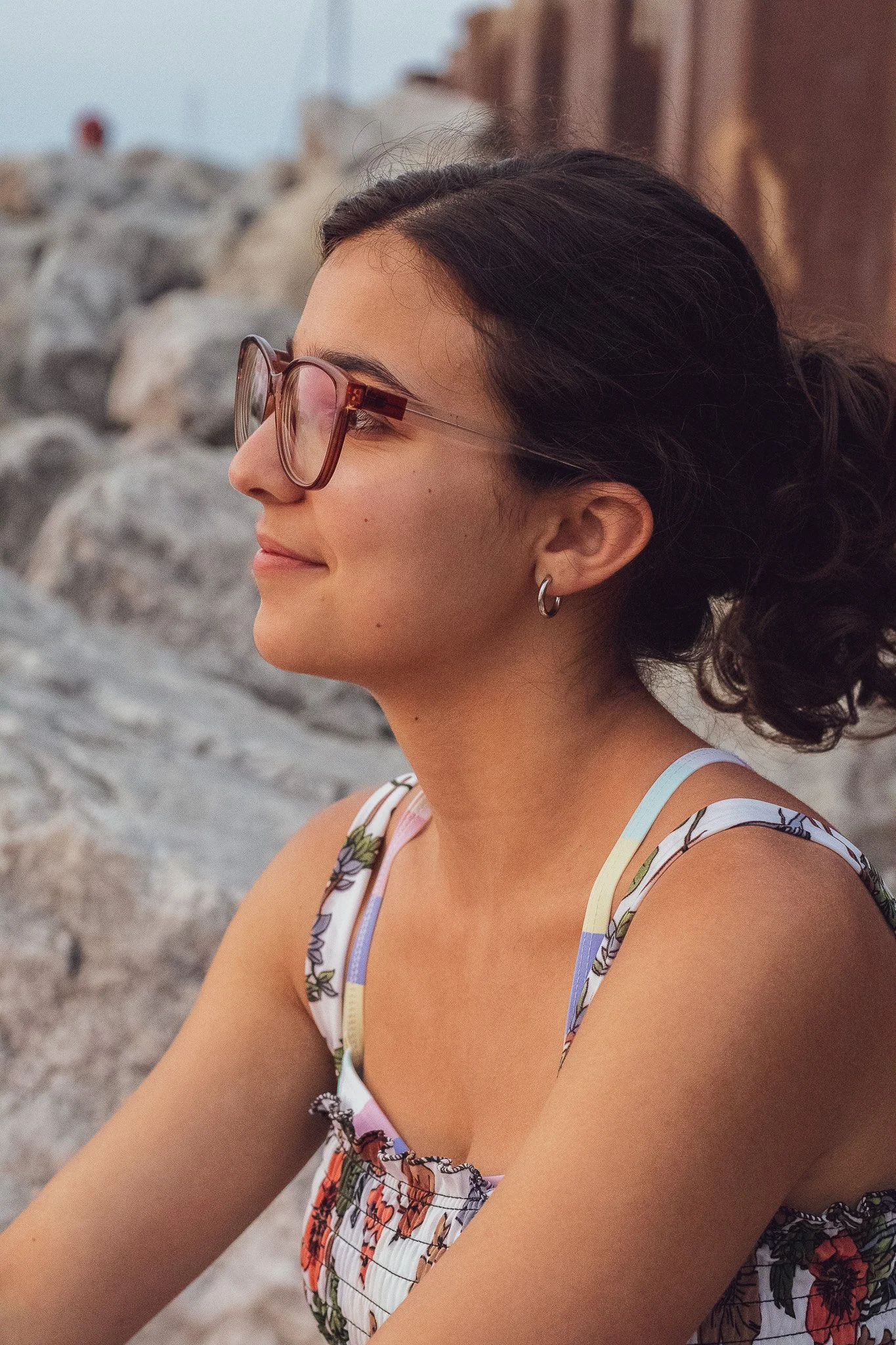 Profile of a young woman with dark curly hair wearing large sunglasses and small hoop earrings. She is smiling slightly and wearing a floral patterned tank top. The background features a rocky shoreline.