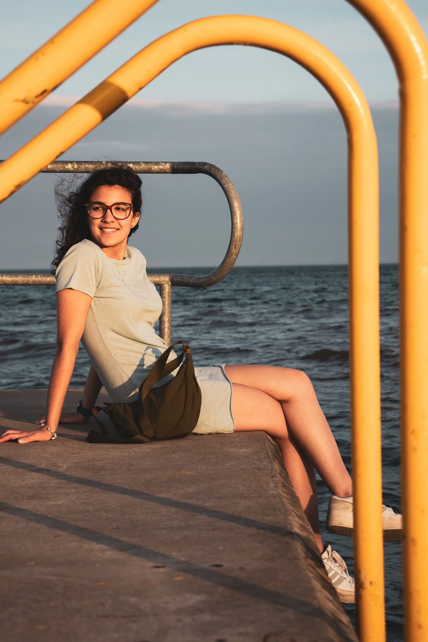 A woman with curly hair and glasses sitting on a dock near the water, smiling, with yellow metal railing and ocean in the background.
