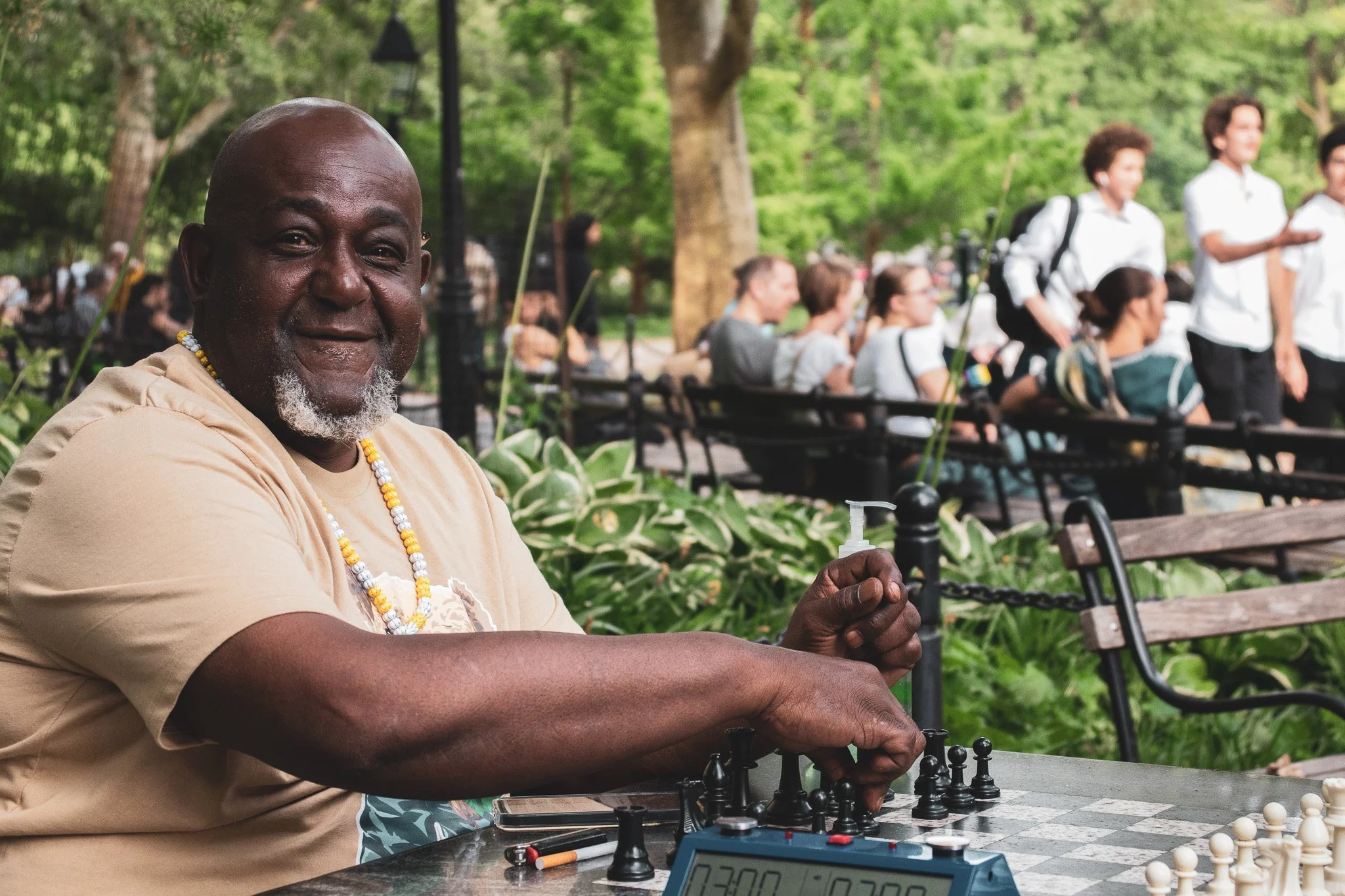 An older man smiling plays chess outdoors in a park, with a chess clock nearby, while other people are seated and standing in the background under green trees.