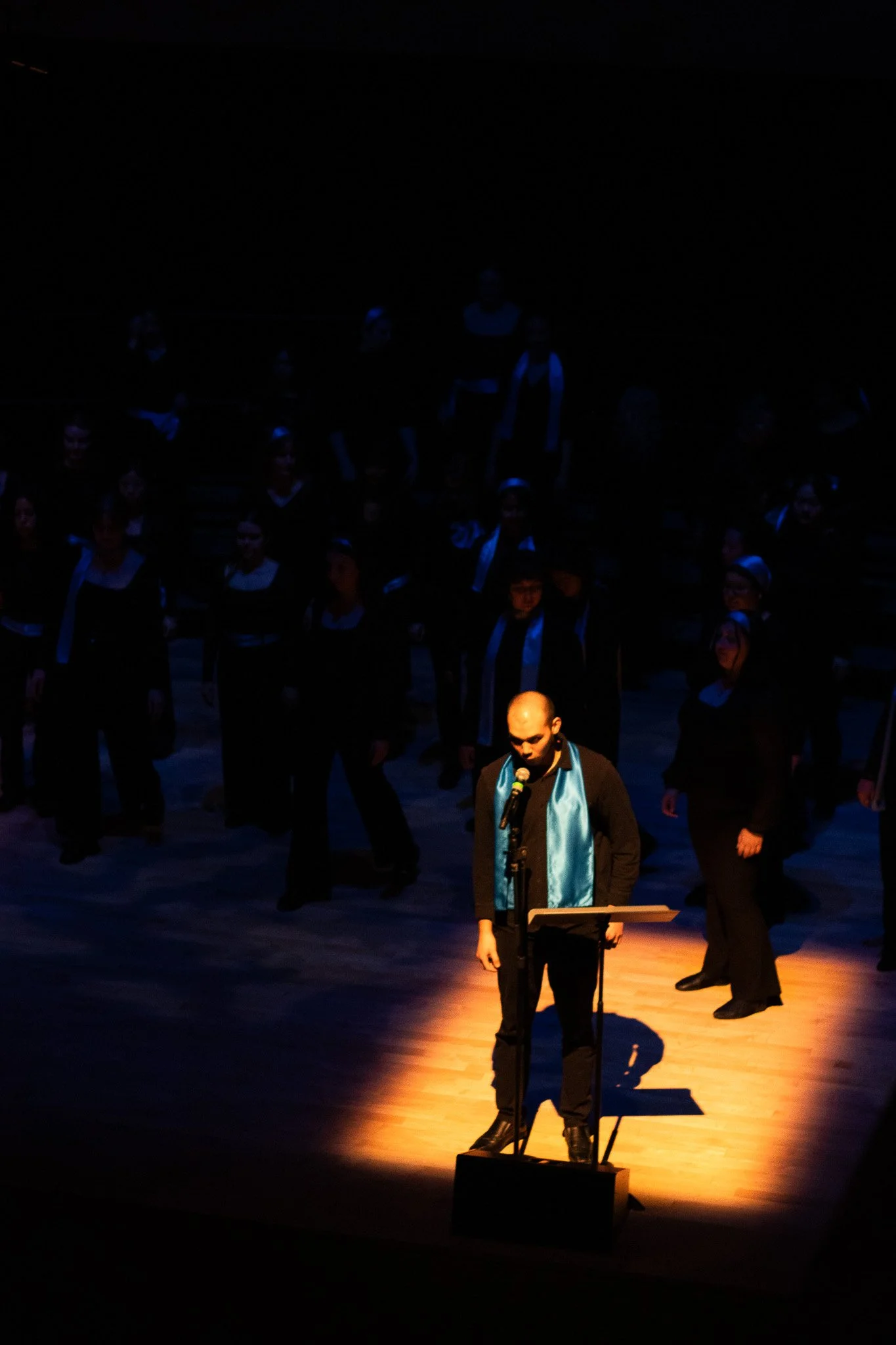 A man in a black suit and blue tie stands at a microphone on a stage with a wooden floor, illuminated by a spotlight, while a choir dressed in black with white collars stands behind him. The background is dark.