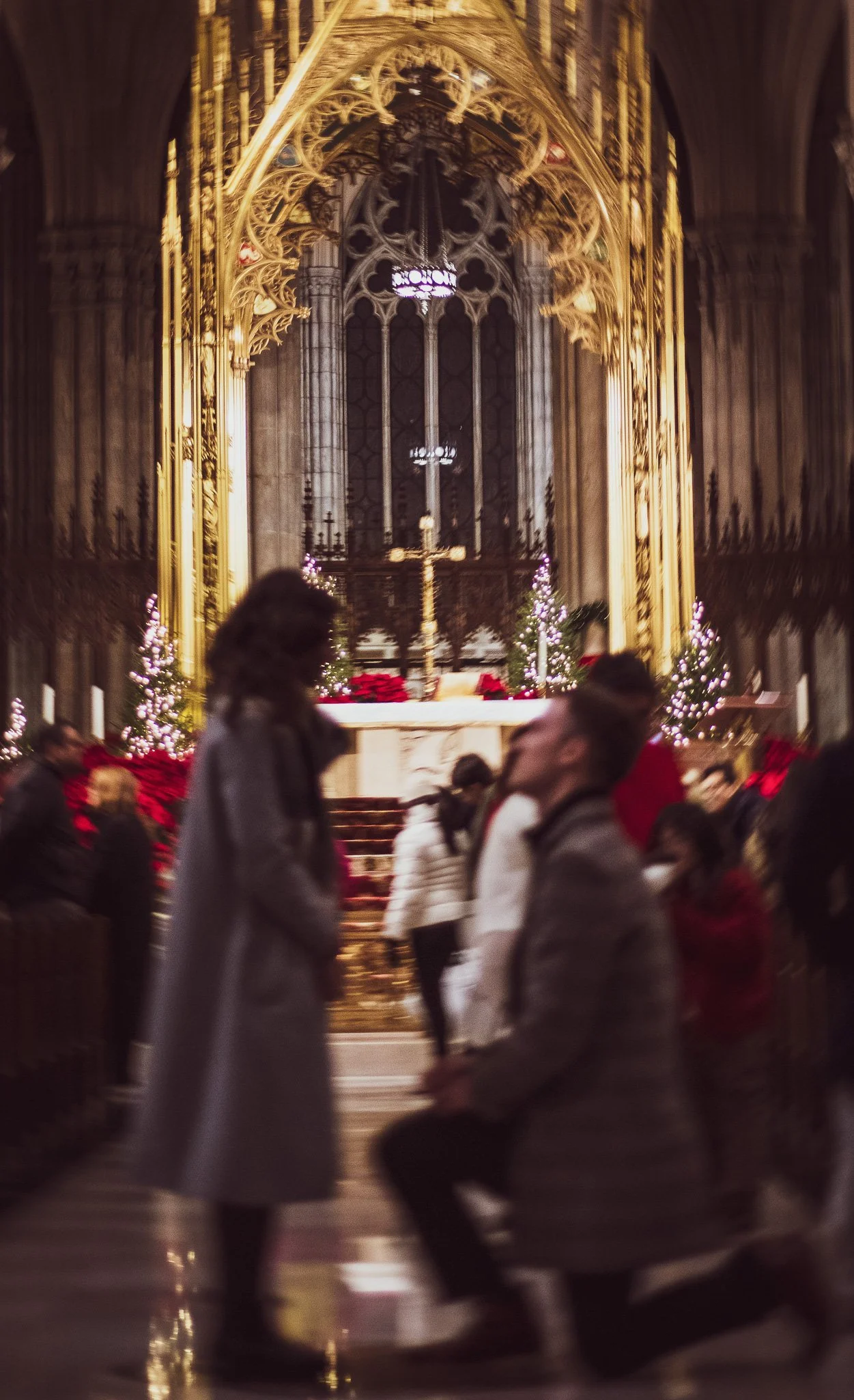 People walking inside a church decorated for Christmas, with small Christmas trees, poinsettias, and ornate golden architecture in the background.