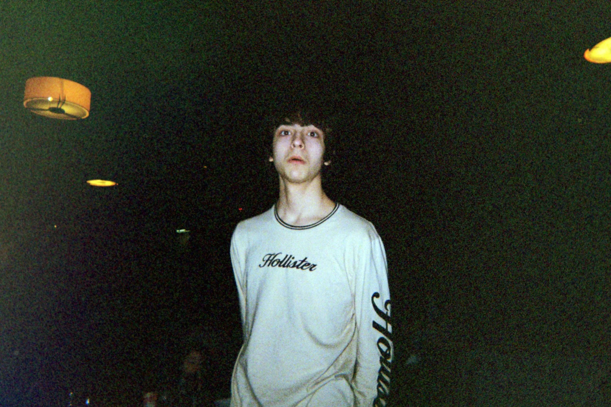A young man with dark hair and a light-colored Hollister t-shirt stands in a dark room illuminated by warm ceiling lights. The background is mostly black.