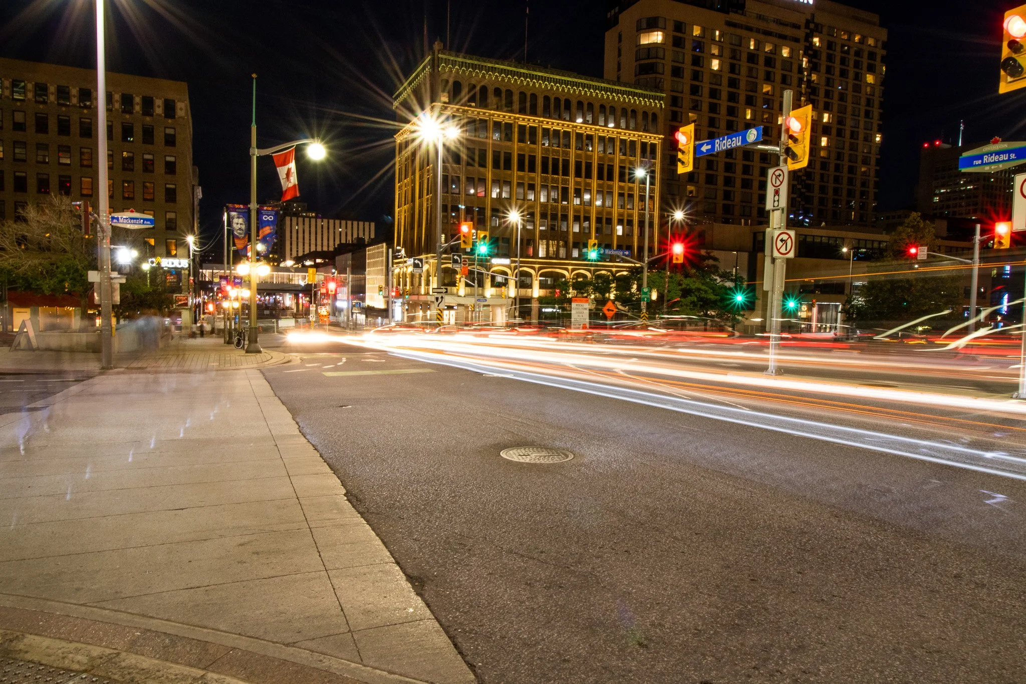 City street at night with illuminated buildings, traffic lights, street signs, and moving vehicle light trails.