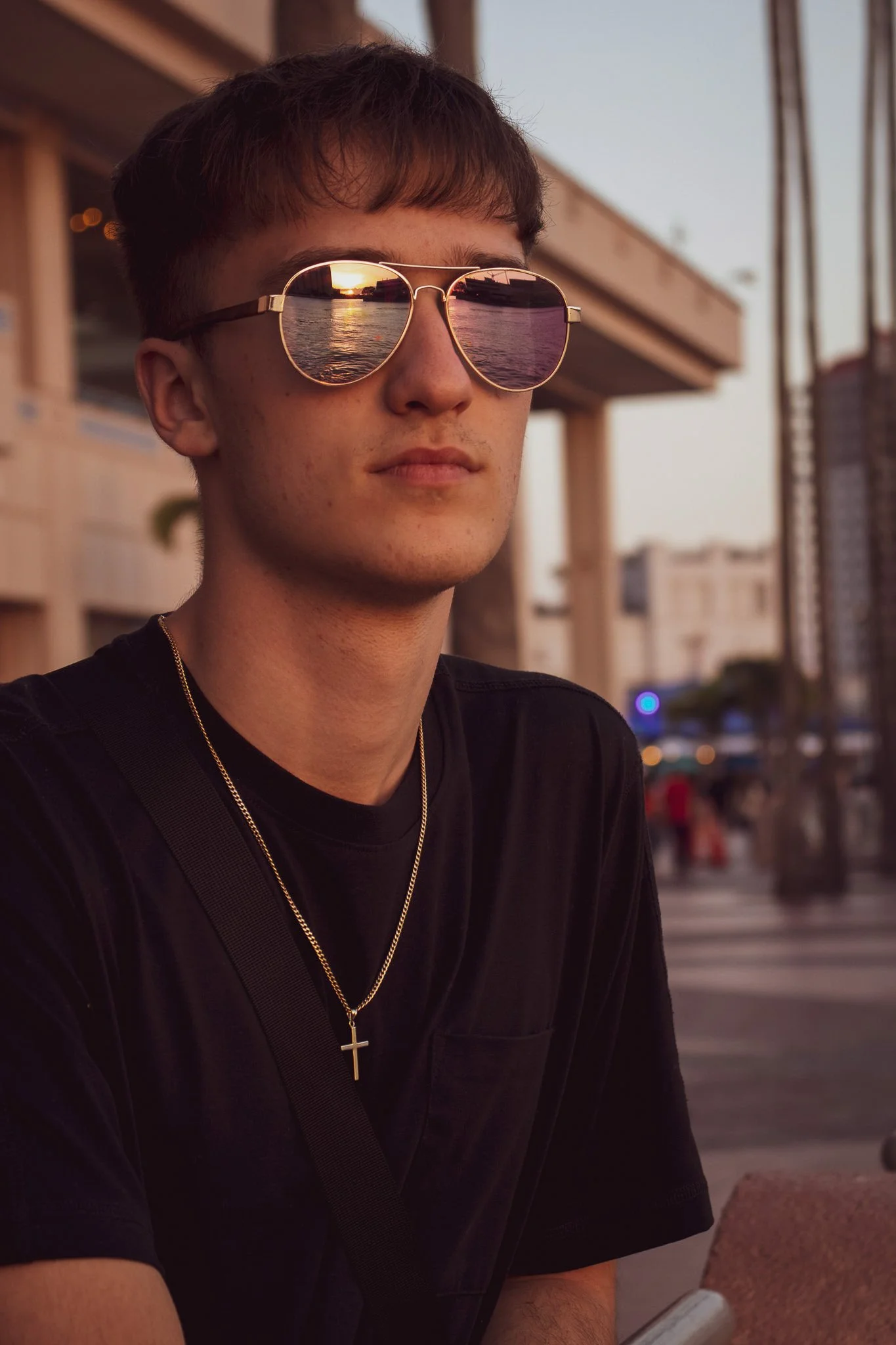 A young man with short brown hair wearing aviator sunglasses reflects a sunset over water. He is dressed in a black shirt with a gold chain and cross necklace, sitting outdoors near a building and street in the evening.