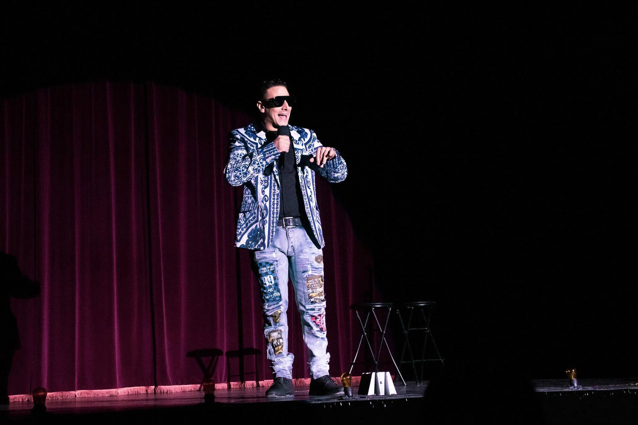 A man wearing sunglasses and a patterned blazer speaking into a microphone on a stage with dark background and purple curtain.