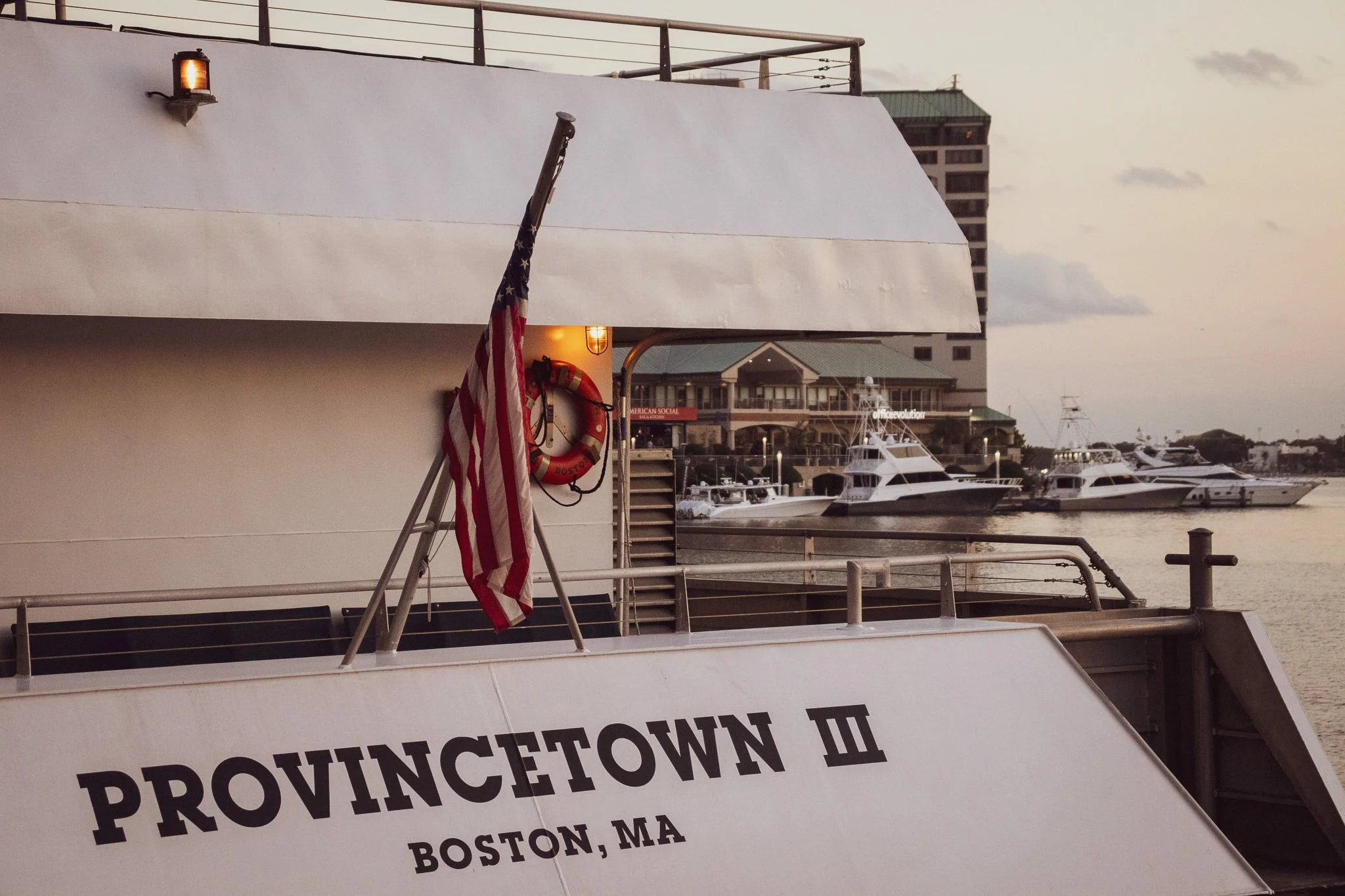 Boat with American flag and life ring docked at a marina, with buildings and yachts in the background during sunset.