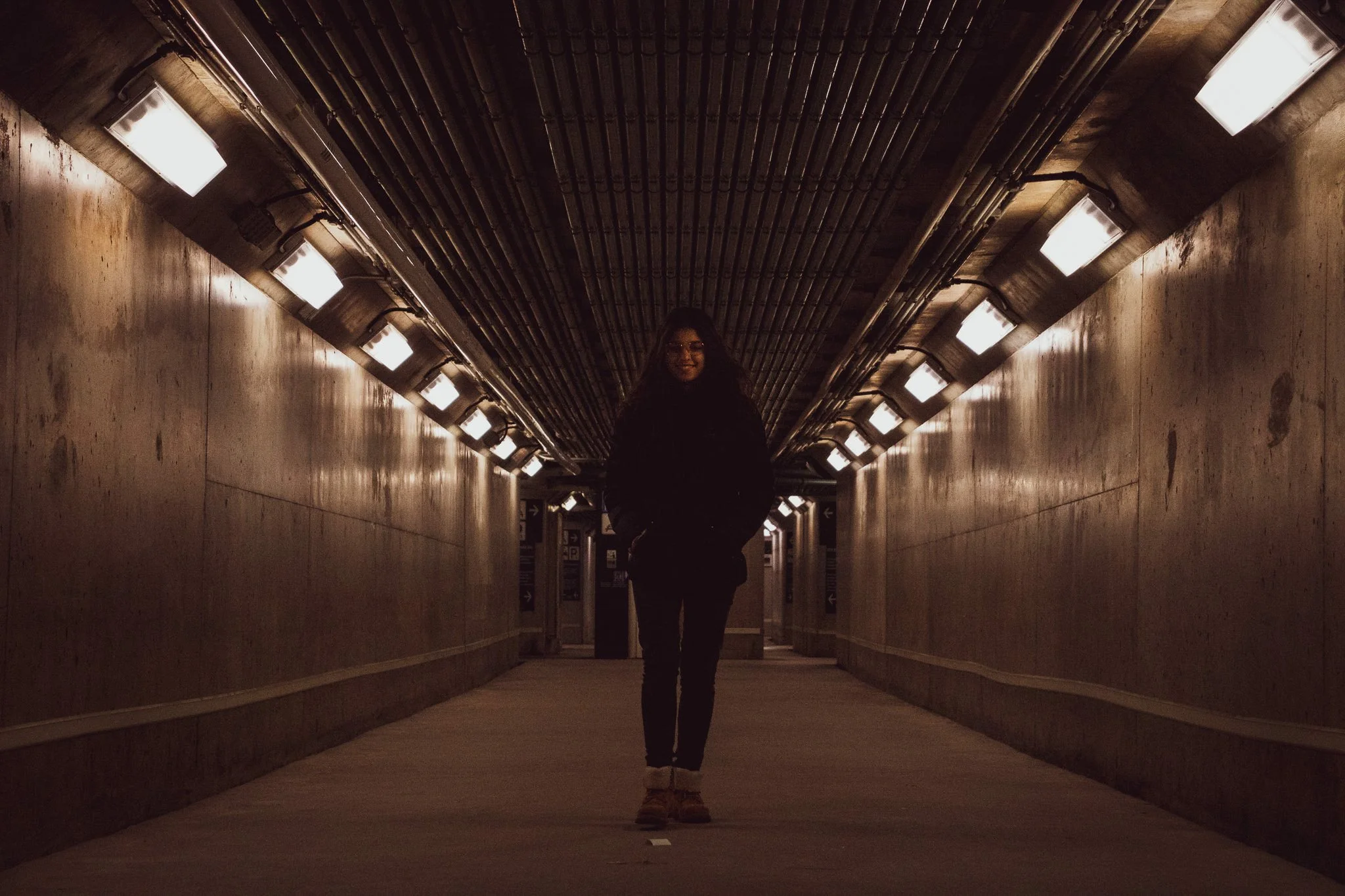 A person standing in a dimly lit underground tunnel with concrete walls and overhead lighting.