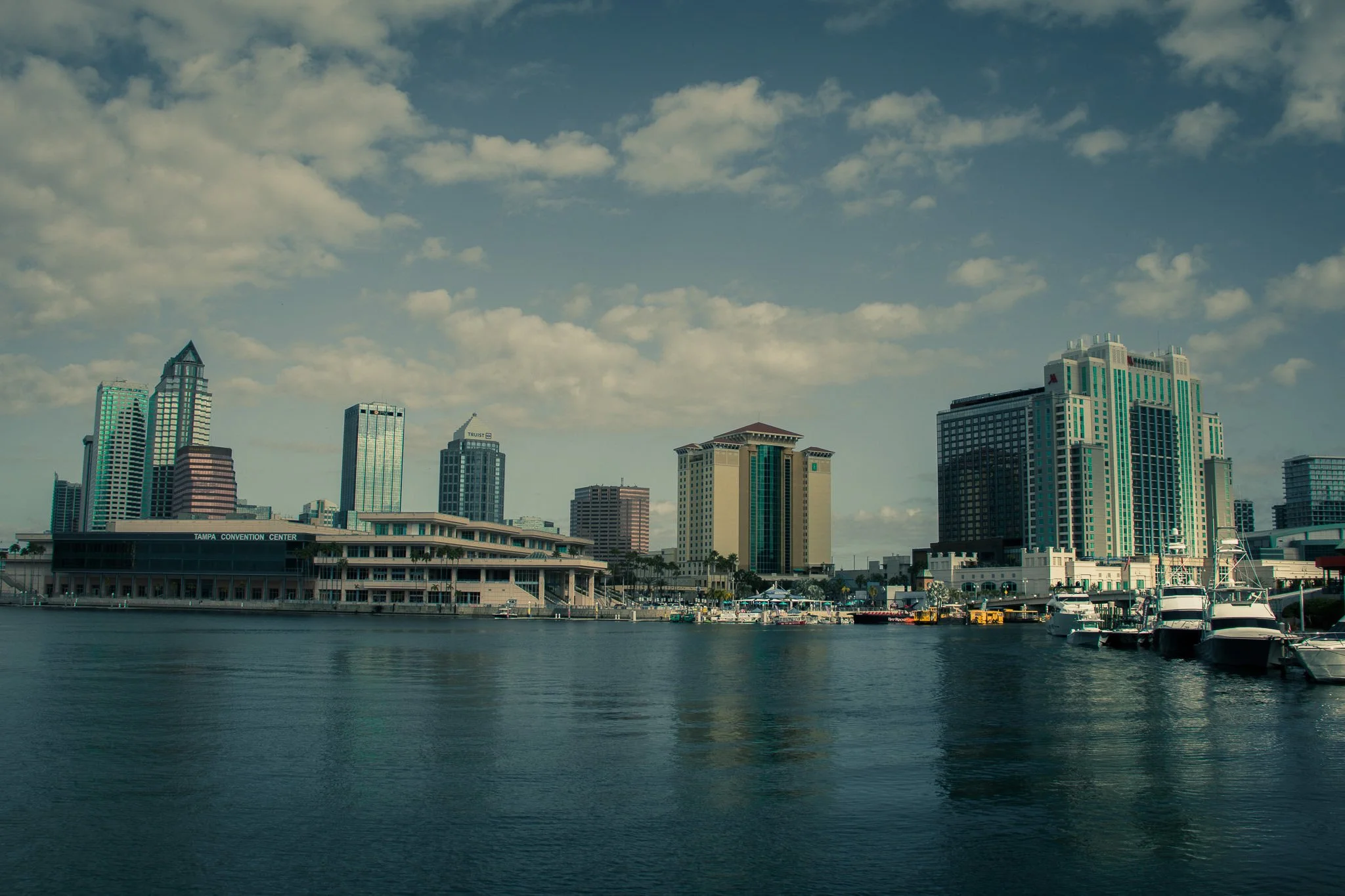City skyline with tall buildings near a waterway, some boats docked, and a partly cloudy sky.