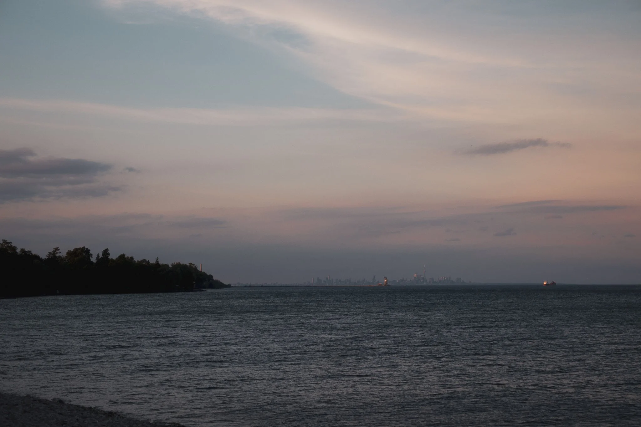 View of a large body of water with a distant skyline and ships, under a sky filled with soft pastel clouds at sunset or sunrise.