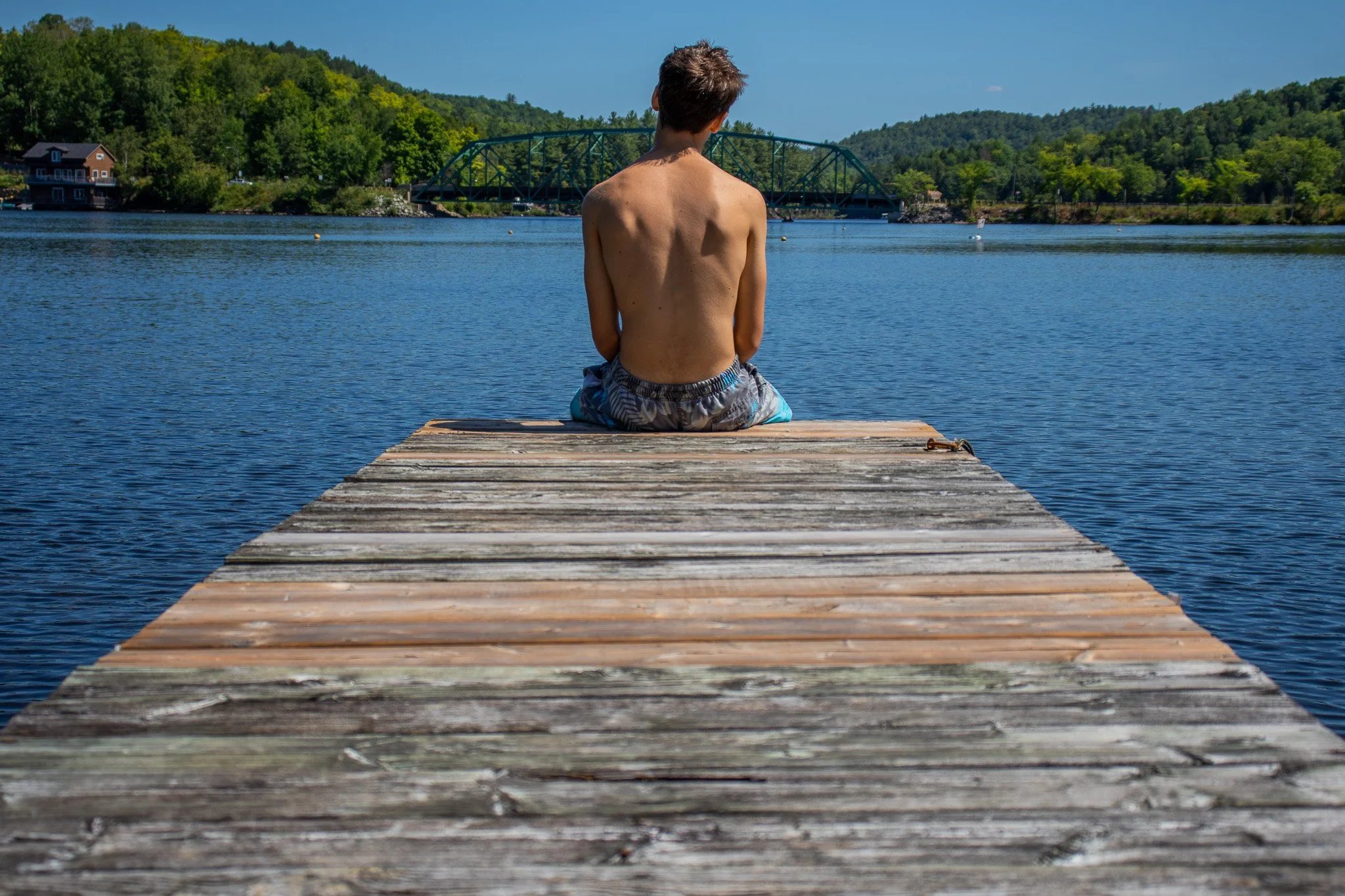 A shirtless young man sitting on a wooden dock by a lake, facing away from the camera, with green trees and houses in the background under a clear blue sky.