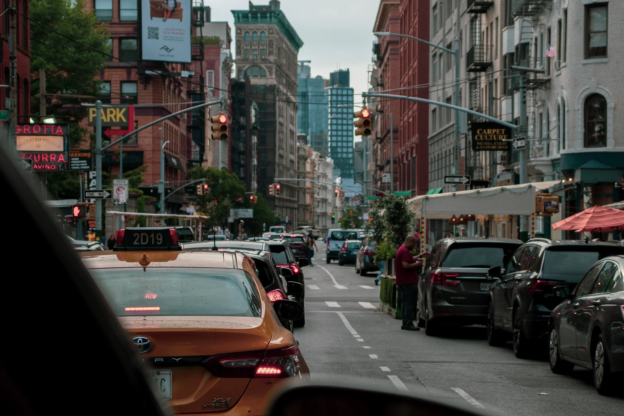 City street view with parked cars, pedestrians, traffic lights, and mid-rise buildings with signs and billboards, under a cloudy sky.