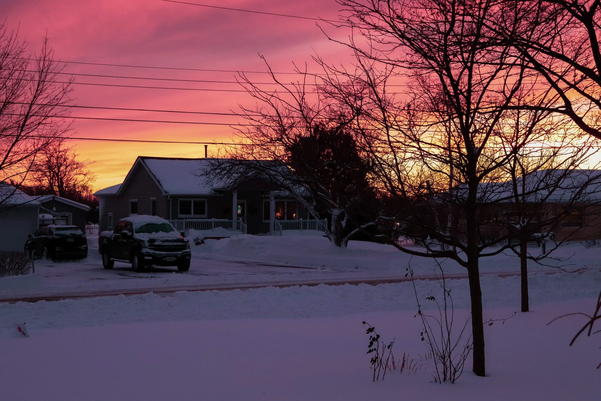 Snow-covered suburban neighborhood during sunset with pink sky and leafless trees, houses, parked cars, and power lines.
