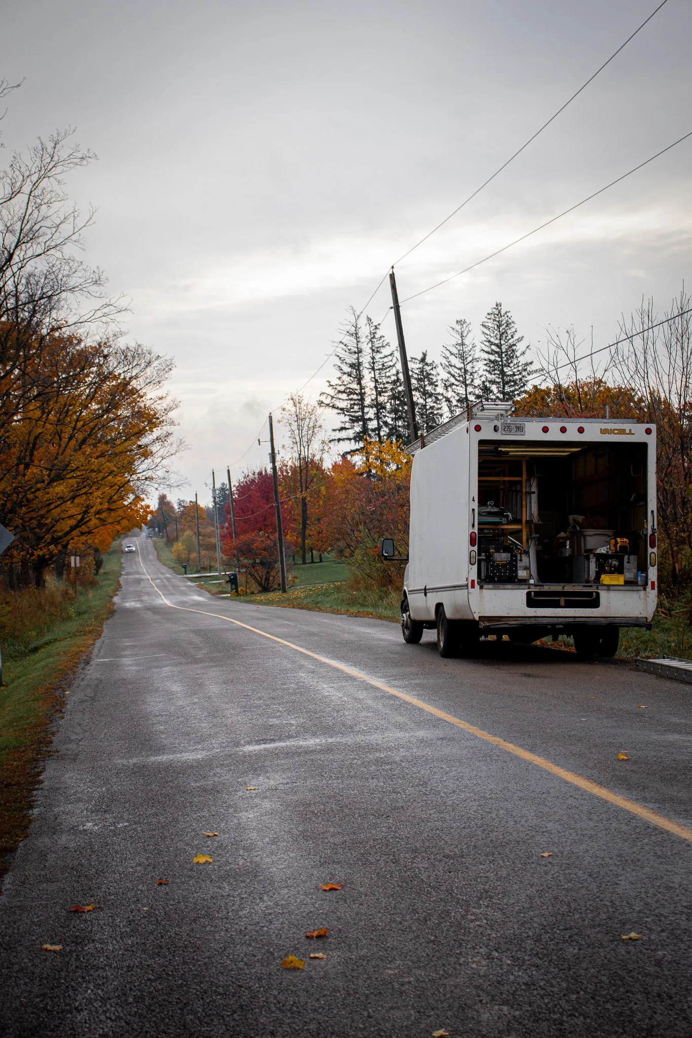 A white moving truck parked on the side of a wet country road during autumn, with colorful trees and a cloudy sky in the background.