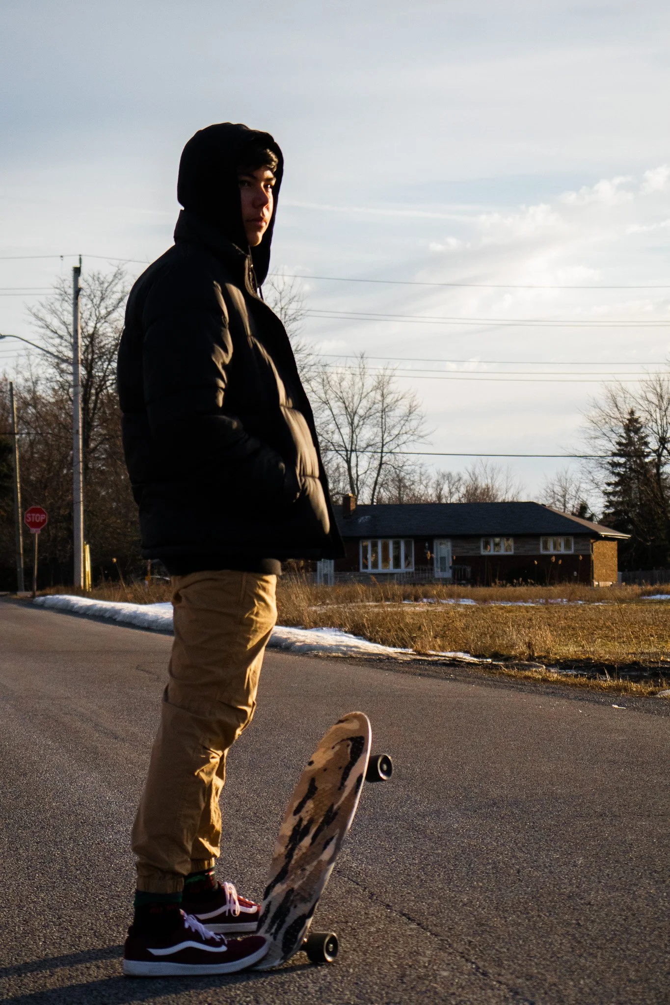 Young person in dark hoodie and tan pants standing on skateboard in neighborhood street during late afternoon, with house, trees, and power lines in background.