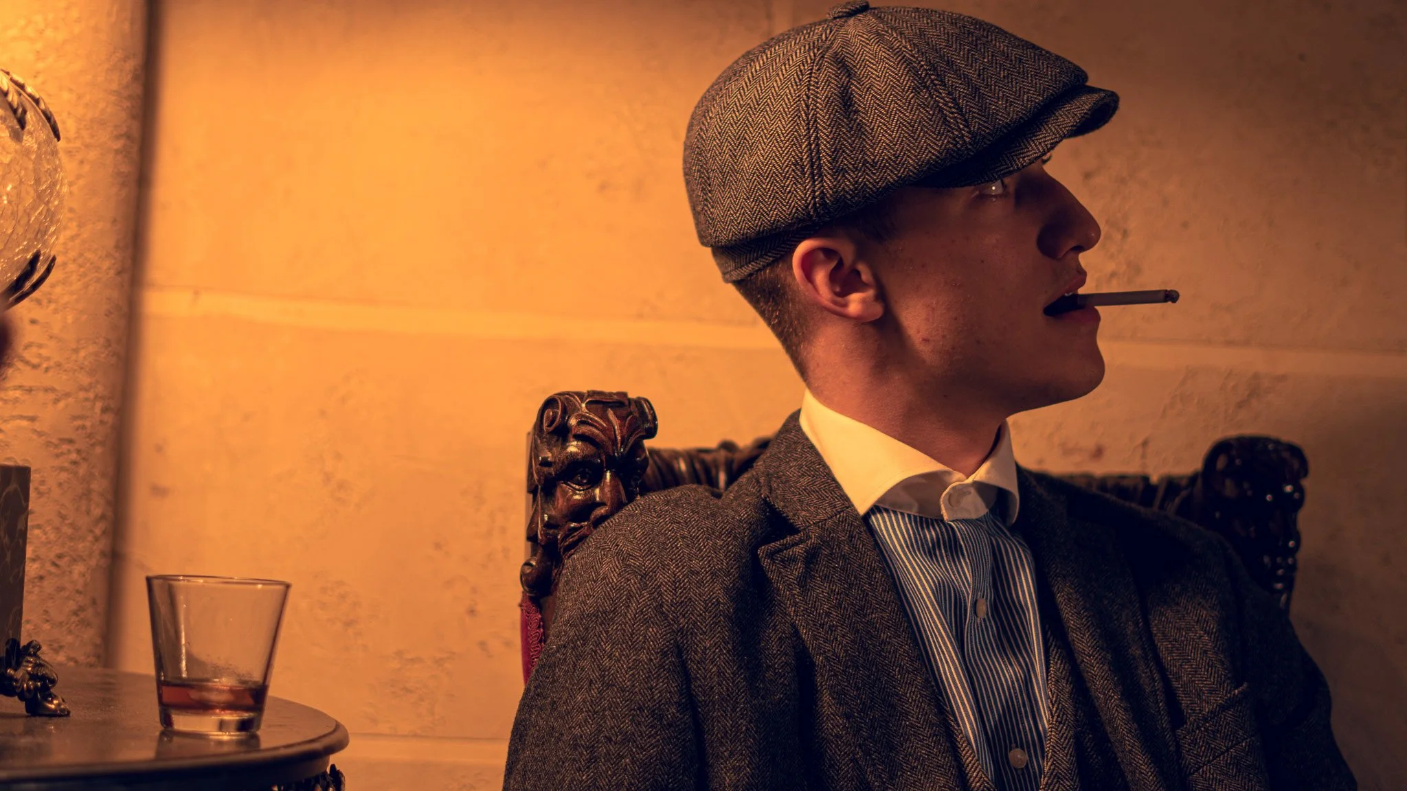 A man with a cigarette in his mouth, wearing a tweed cap and a vintage suit, sitting in a dimly lit room.