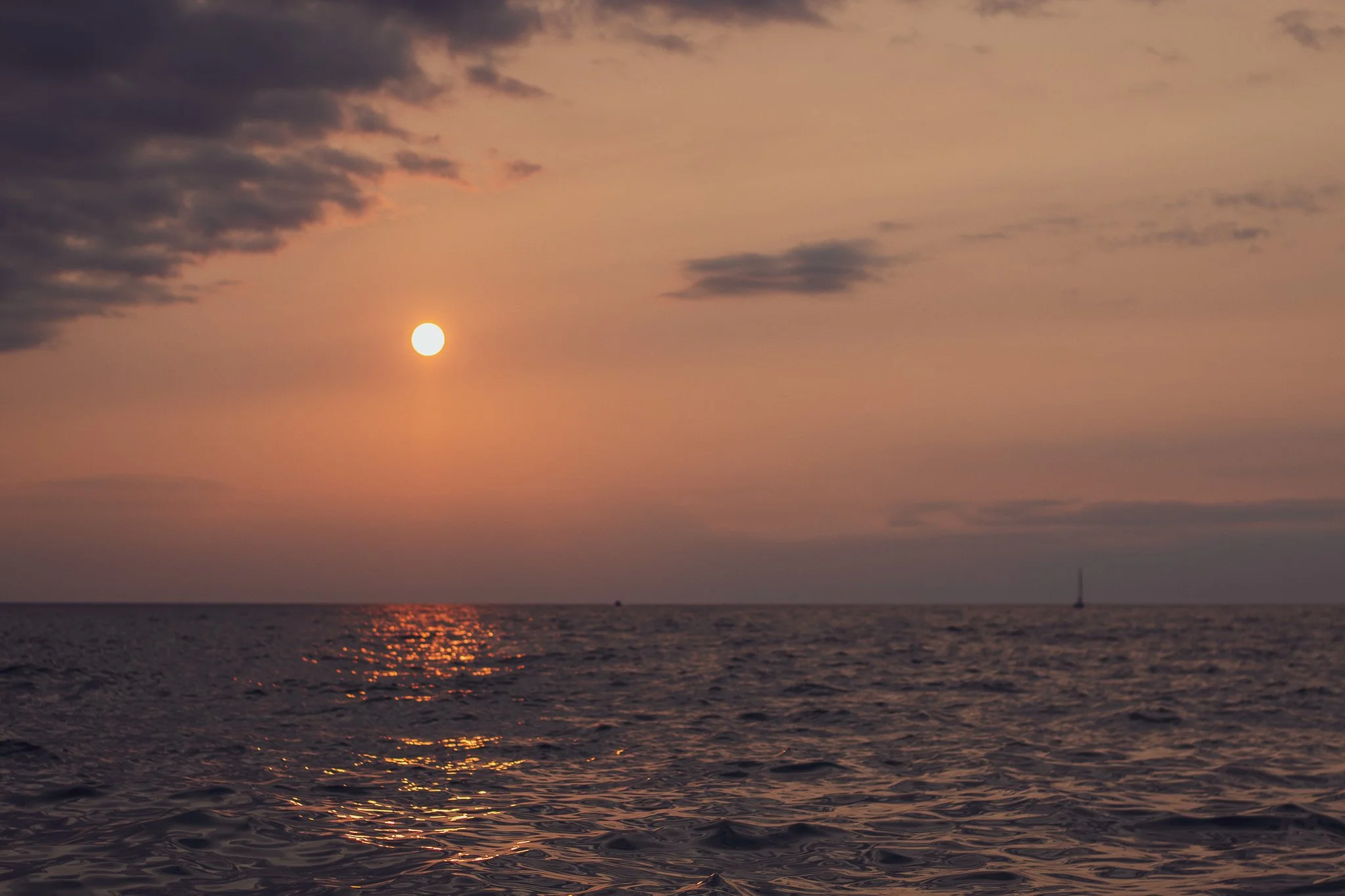 Sunset over the ocean with a sailboat in the distance, clouds in the sky, and the water reflecting the warm colors of the sunset.