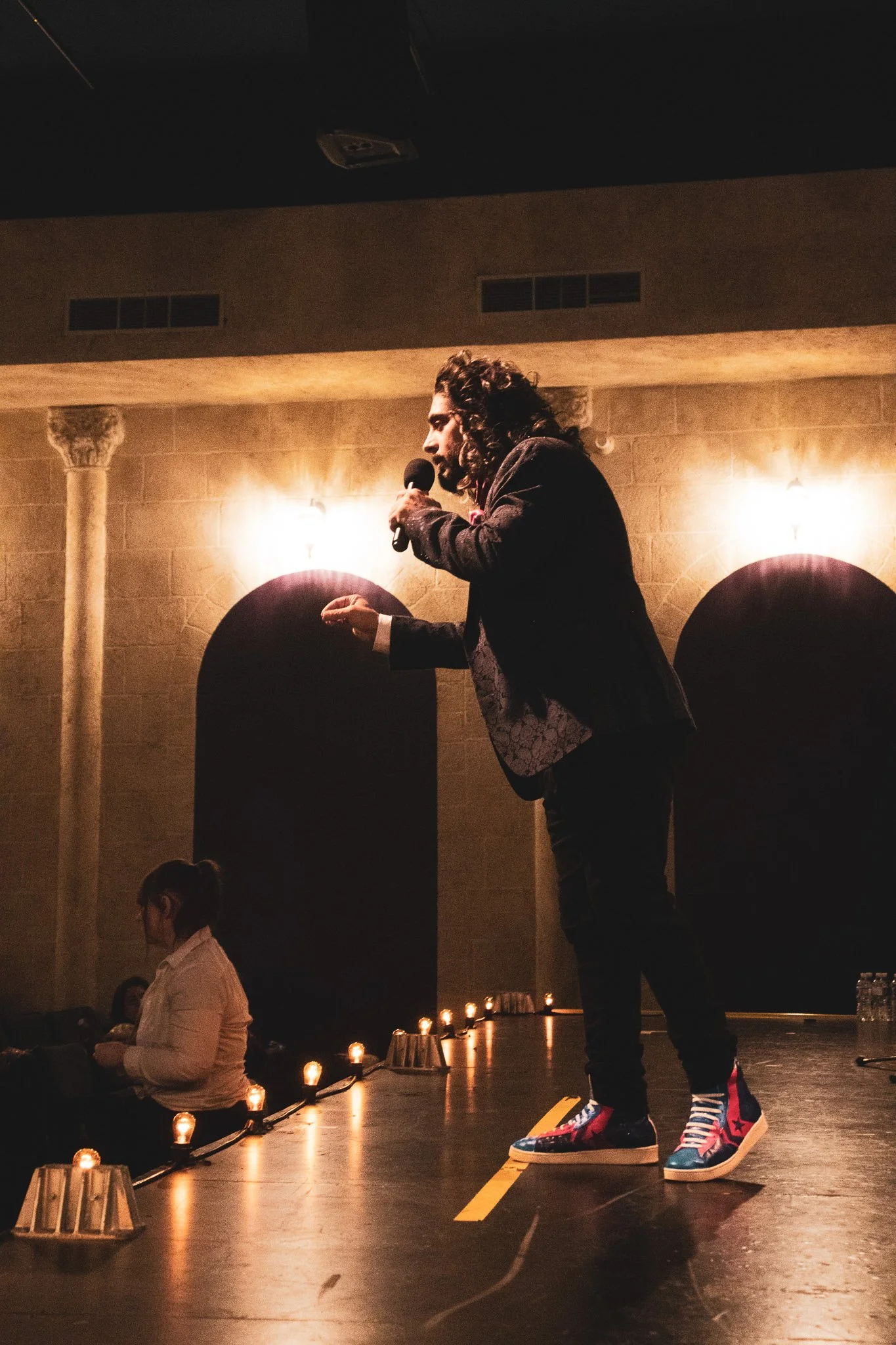 A man with curly hair wearing colorful sneakers is standing on a skateboard on stage, holding a microphone and speaking. There are small candles lined up on the floor in front of him, and a woman is sitting nearby, looking down. The background has wa