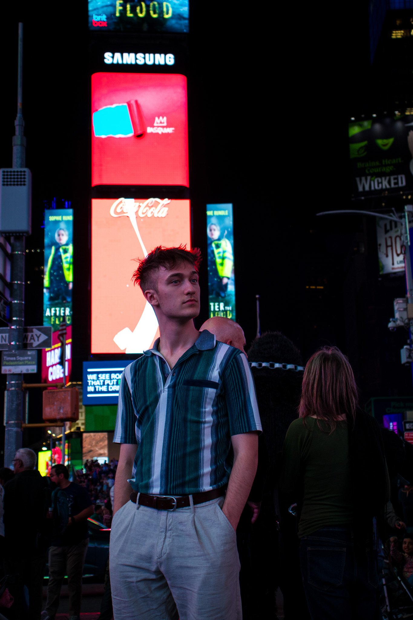 A young man with short hair wearing a striped blue and green shirt standing in Times Square at night, surrounded by illuminated billboards and other people.