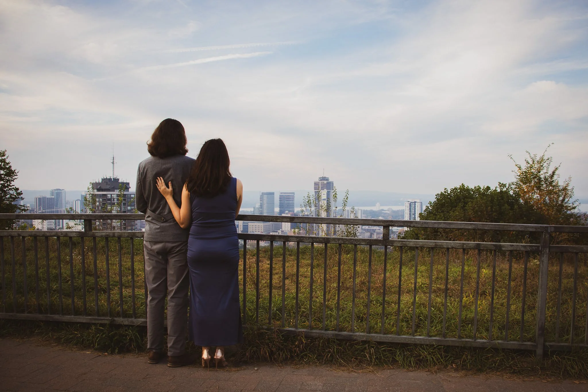 Two women standing side by side at an outdoor viewpoint overlooking a city skyline with tall buildings, trees, and a body of water in the distance.