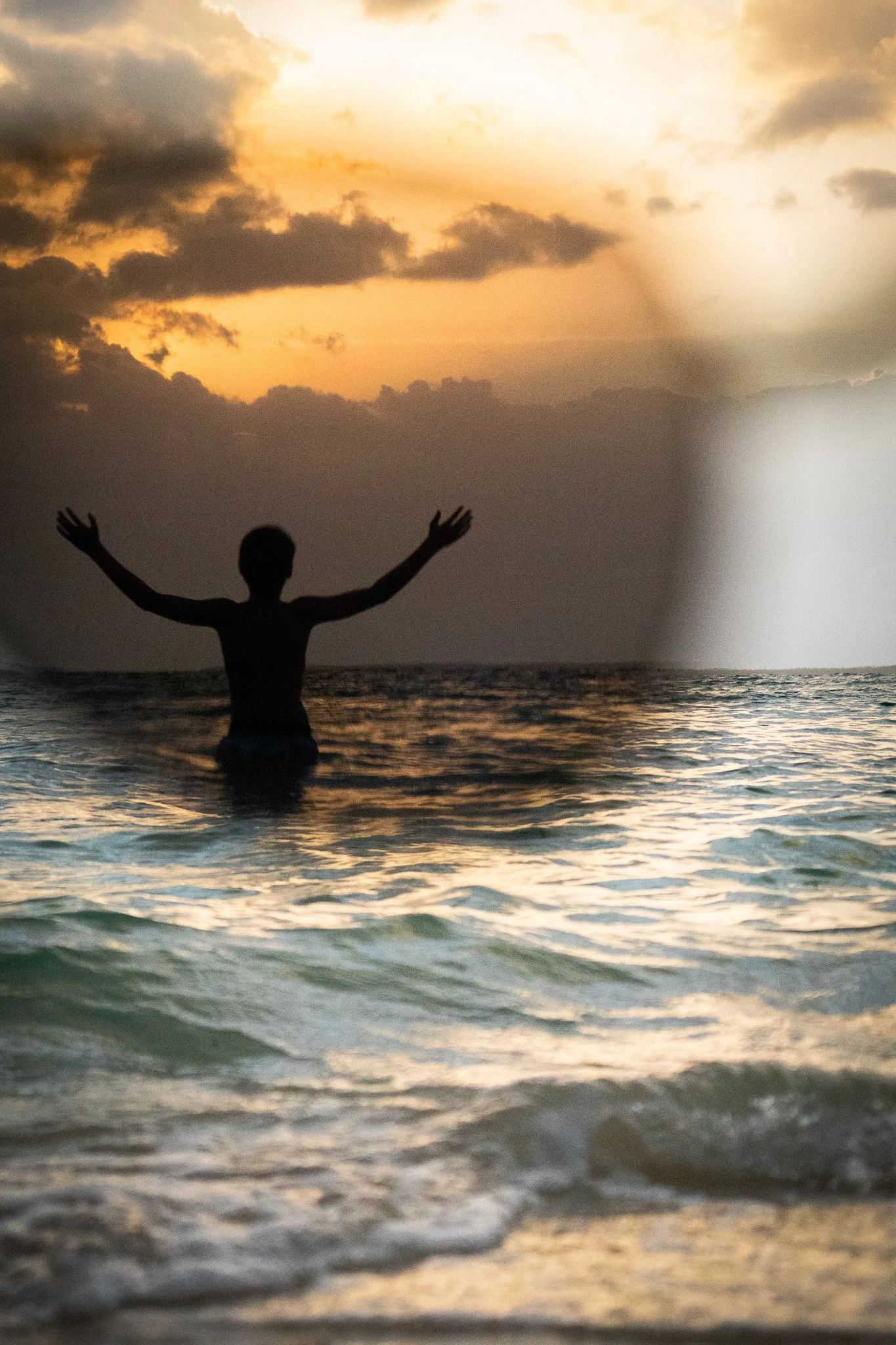 Silhouette of a person with arms raised in the water at sunset, with clouds and a golden sky in the background.
