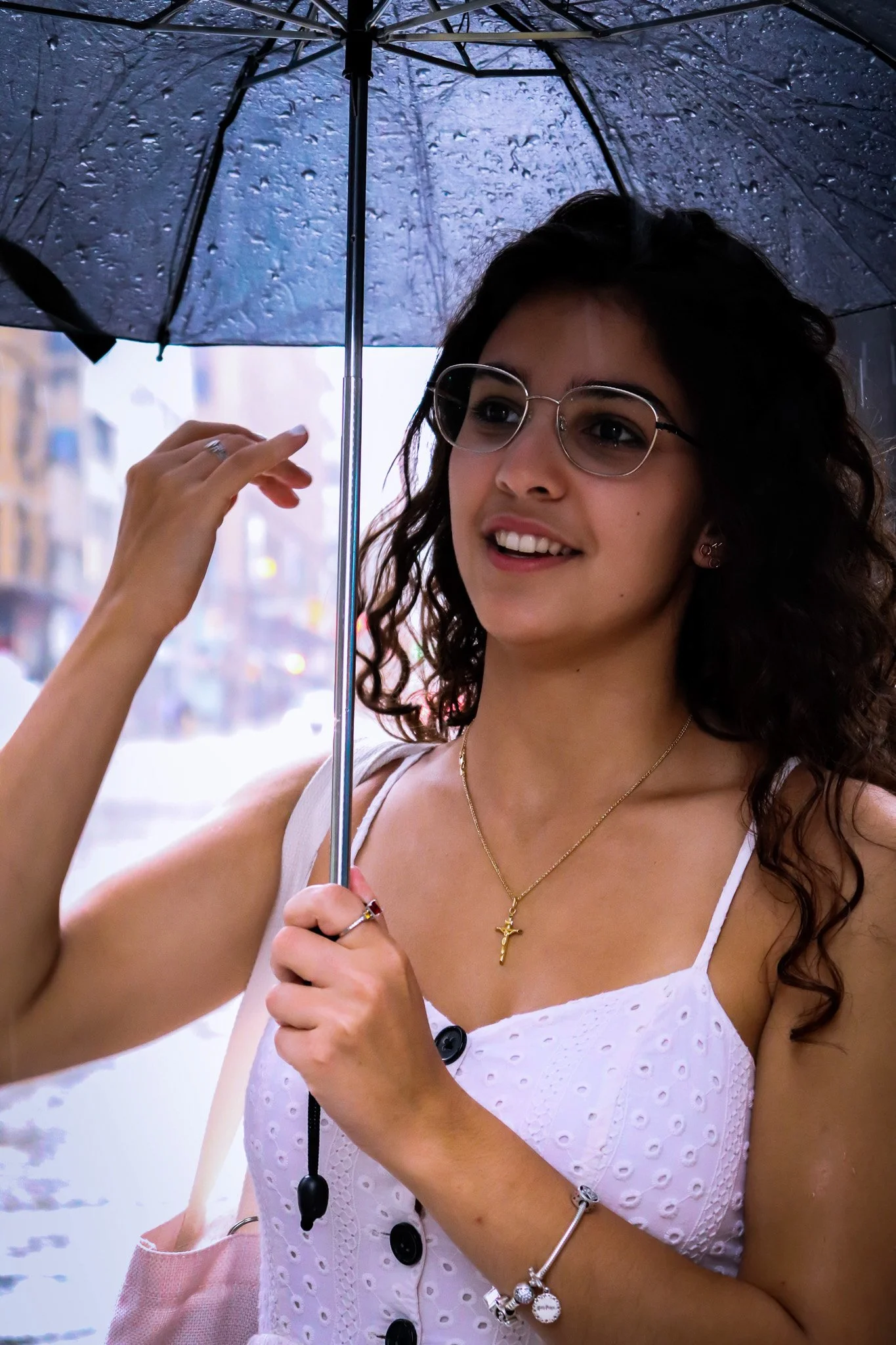 A woman with dark, curly hair wearing glasses, a white sleeveless top, a gold cross necklace, and jewelry, holding a black umbrella on a rainy day in an urban setting.