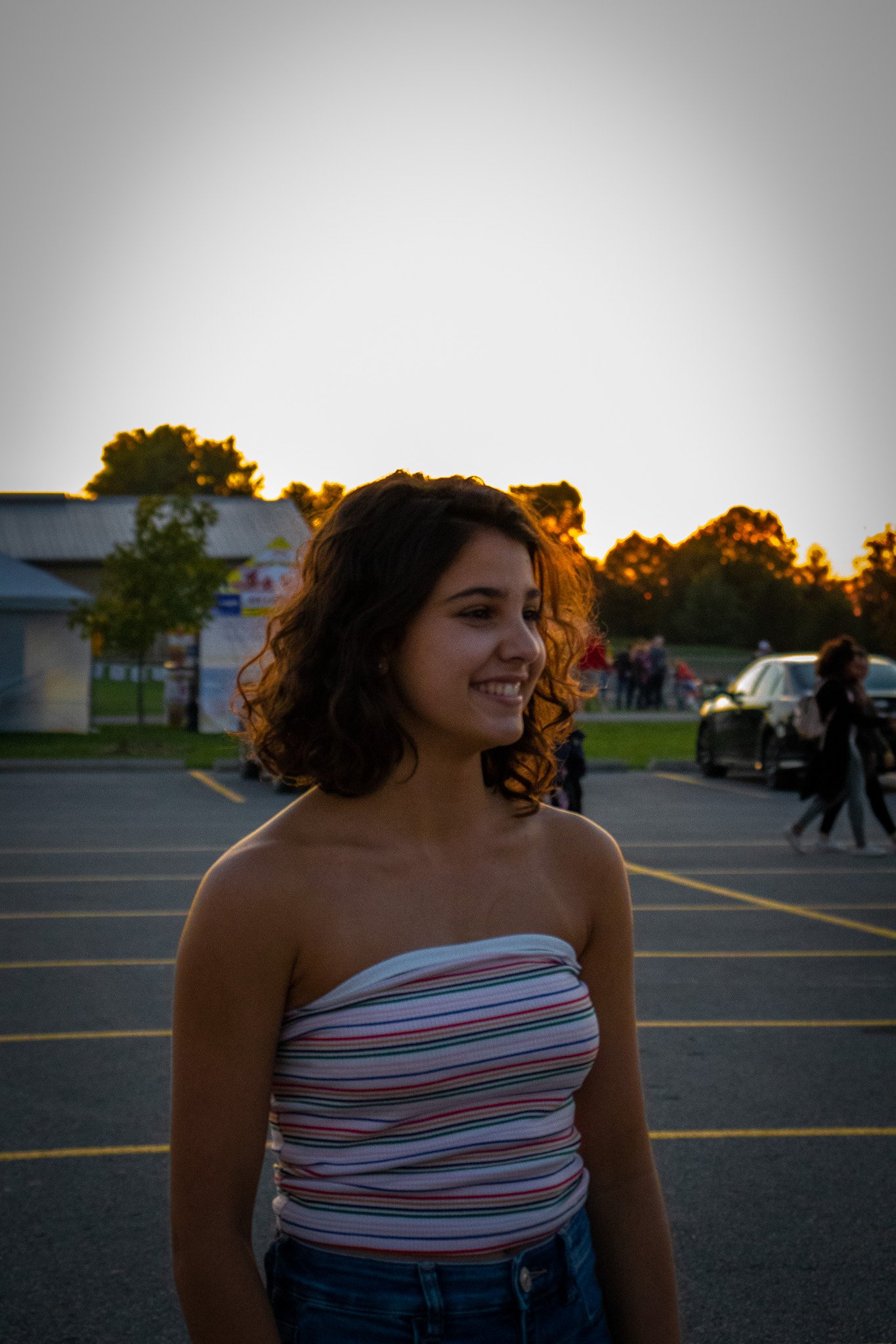 A young woman with curly brown hair smiling, wearing a strapless, striped top, standing in a parking lot at sunset with trees, cars, and people in the background.
