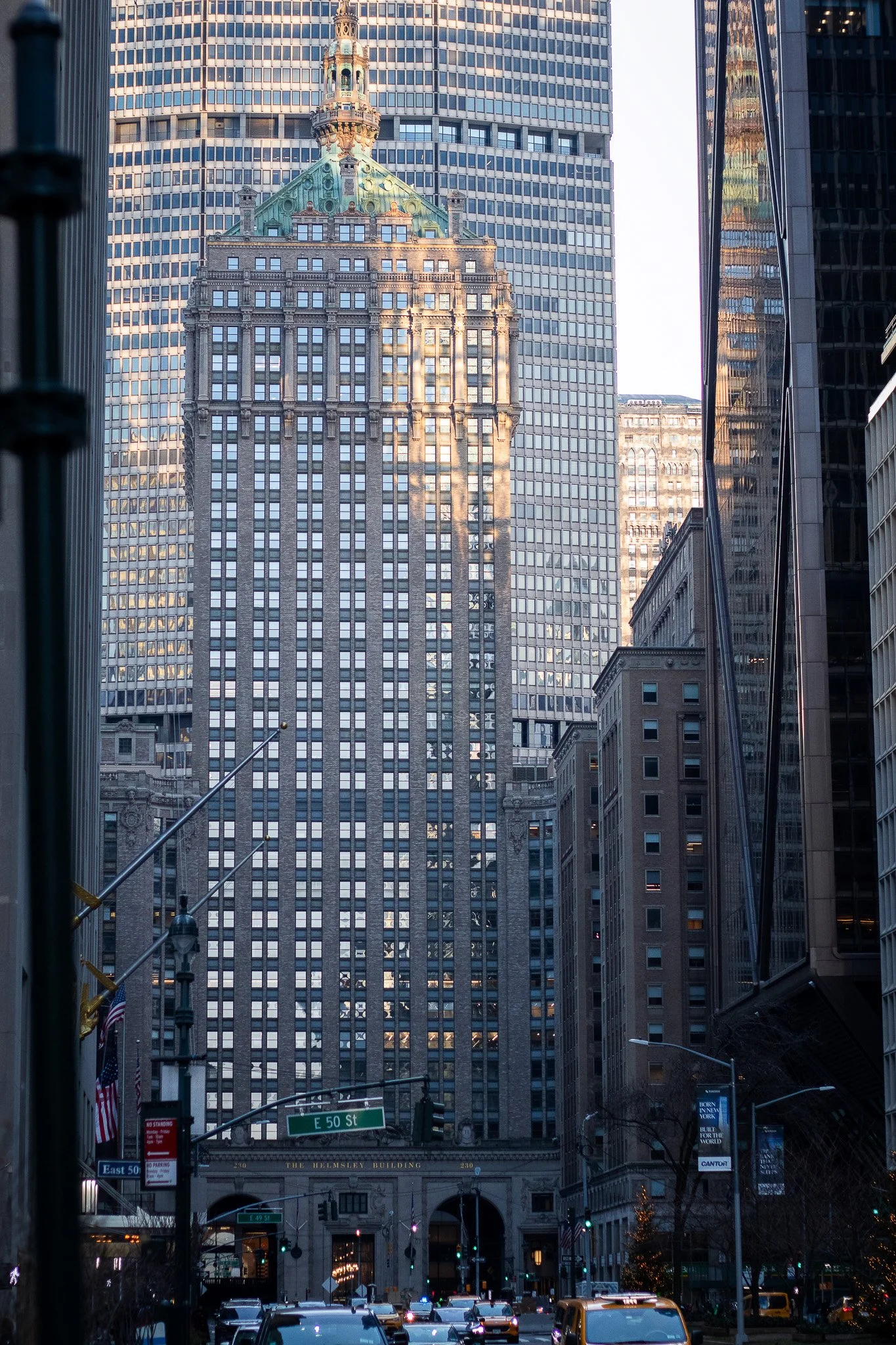 Tall buildings in a cityscape, with one having a green tower and ornate top, framed by other glass skyscrapers. Street signs indicate E 50th St, and cars are visible on the street below.