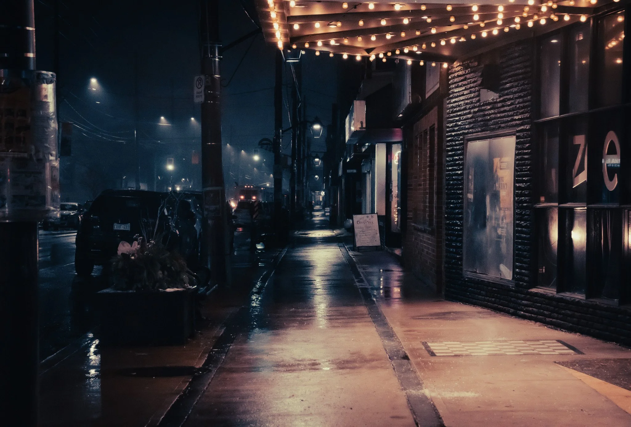 Nighttime scene on a city sidewalk with wet pavement reflecting streetlights and storefront lights, and an awning decorated with string lights.