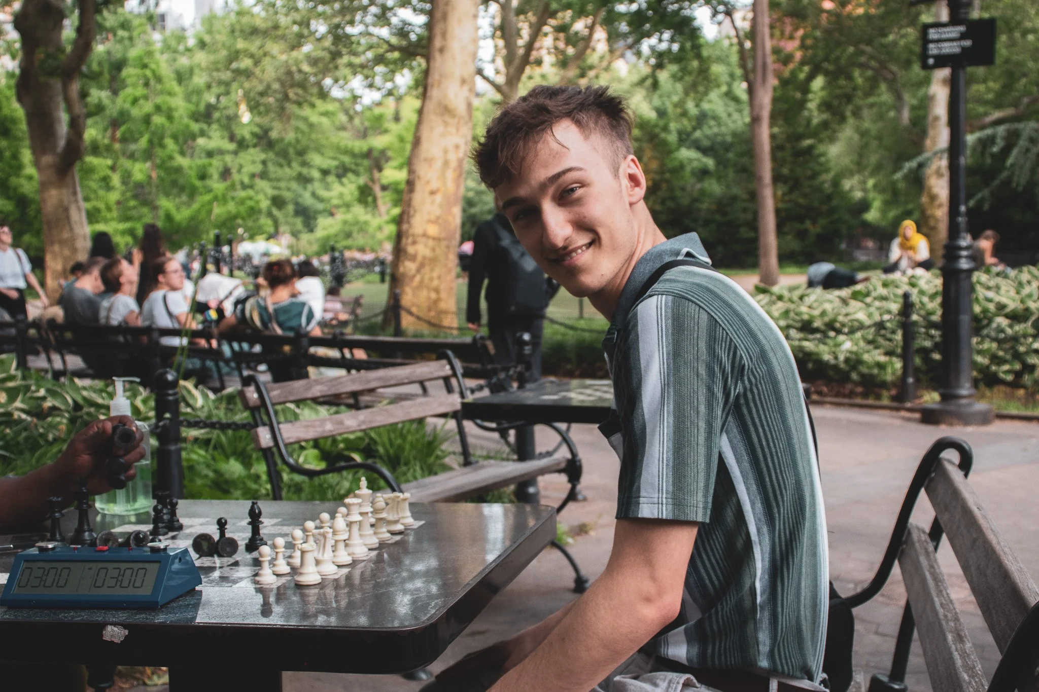 A young man with short, dark hair is playing chess outdoors at a park. He is smiling and Turned to the camera. There are people sitting on benches and trees in the background.